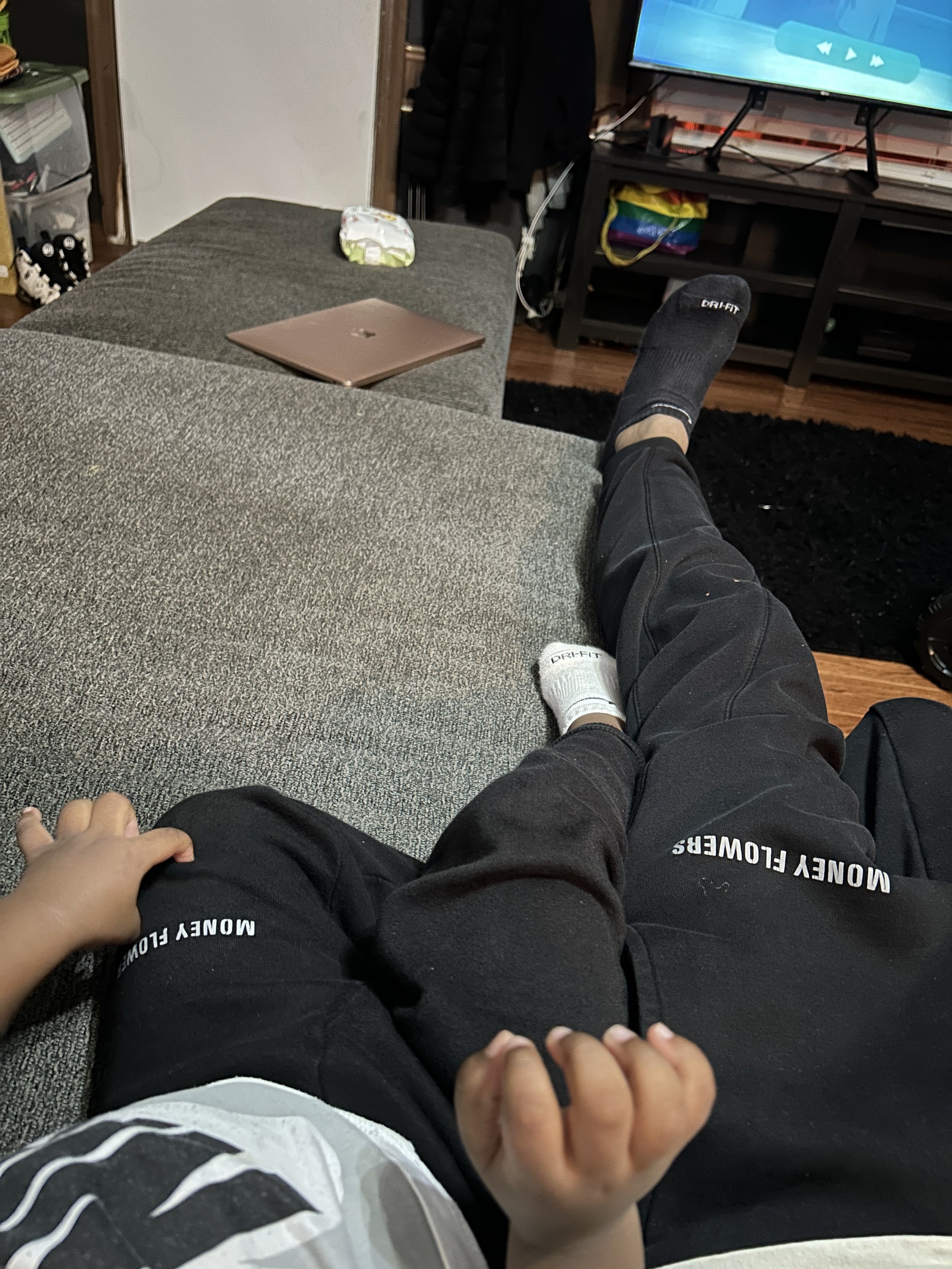 A person is sitting on a gray couch with their legs stretched out. They are wearing black athletic pants with the words 'MONEY FLOWERS' on one pant leg, black socks, and a long sleeve shirt. A child's hand is resting on their thigh, holding onto thei