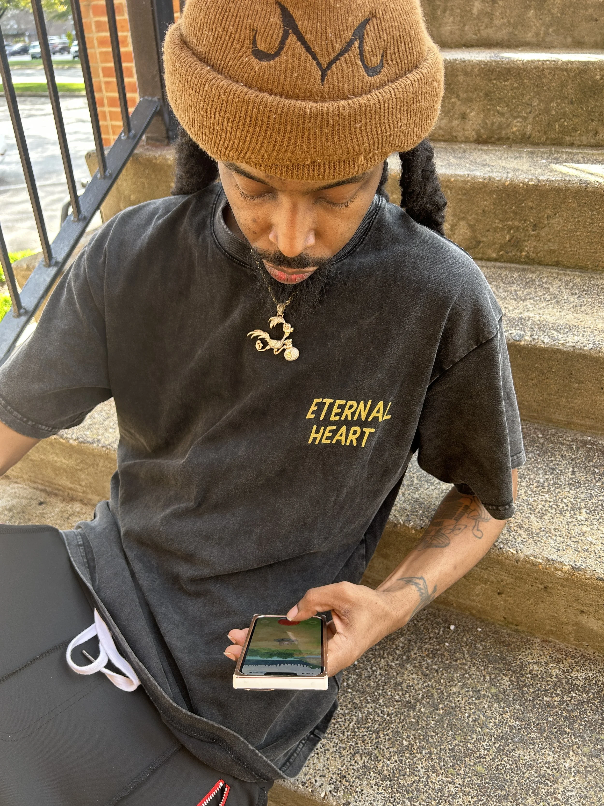 A person sitting on outdoor stairs wearing a brown beanie with a black logo, a black T-shirt with yellow text that says "ETERNAL HEART," a necklace with a dragon pendant, and holding a smartphone while looking down at it.