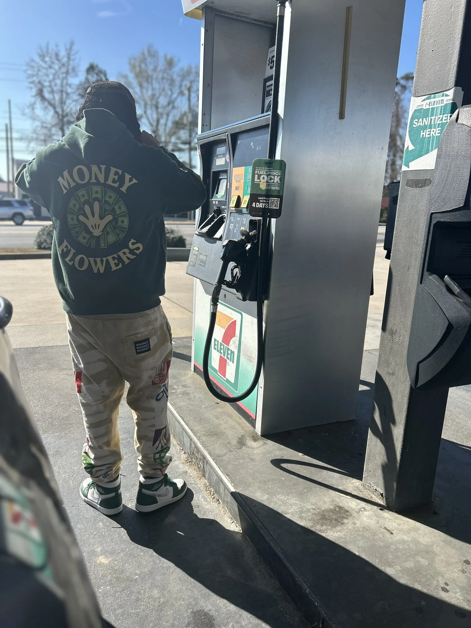 A person wearing a hoodie with 'Money Flowers' and a hand design, and baggy pants with patches and sneakers, standing at a 7-Eleven gas pump on a sunny day.