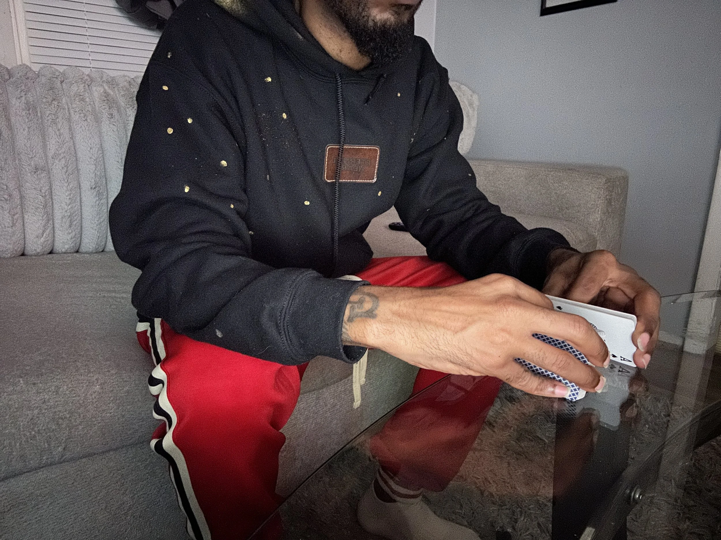 A man sitting on a beige couch, playing with a tablet. He has a black hoodie with paint splatters, red shorts, and tattoos on his arms.
