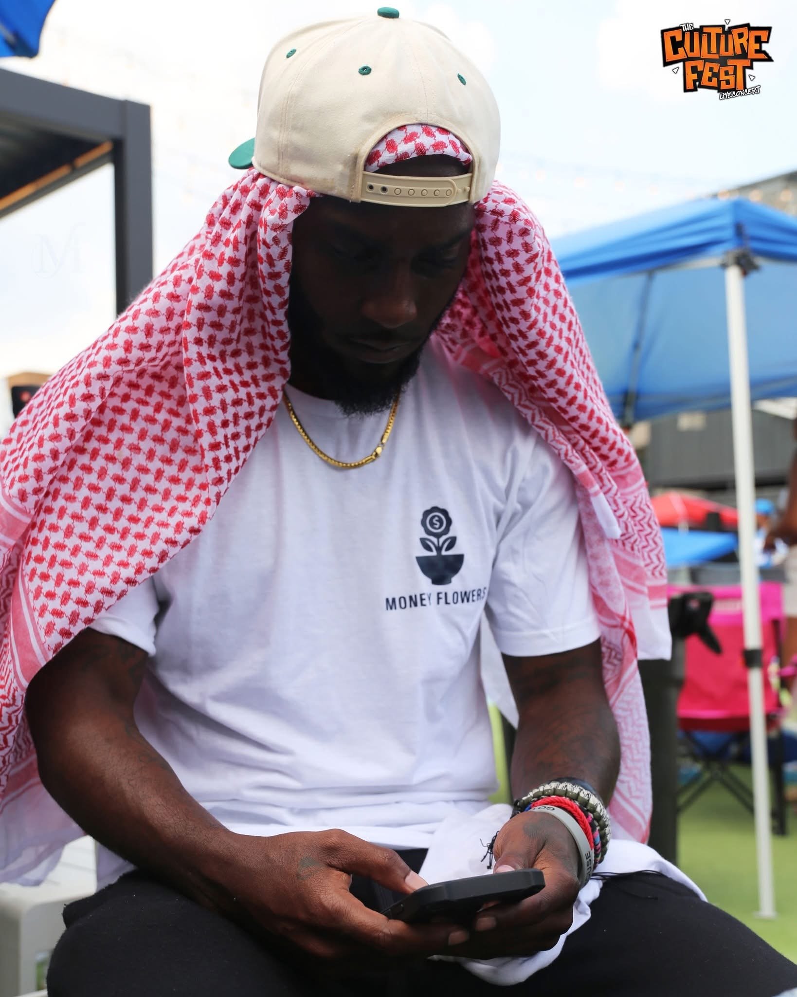A person with dark skin wearing a beige baseball cap backward, a red and white checkered cloth draped over shoulders, a white t-shirt with a black logo, and multiple bracelets, looking at a phone. In the background, there is a blue tent and a logo in