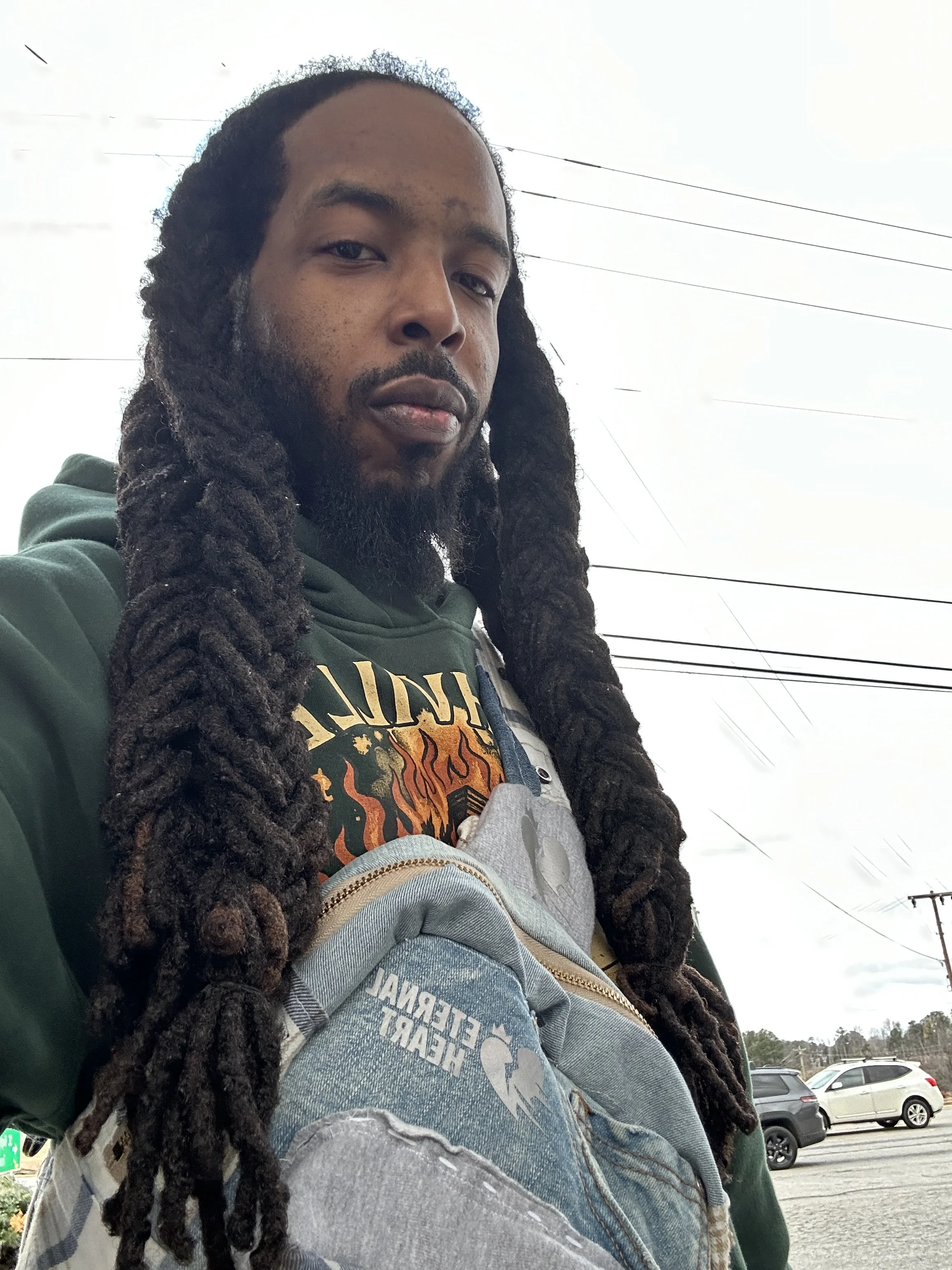 Close-up of a man with long dreadlocks and a beard taking a selfie outdoors, with parked cars and power lines in the background.