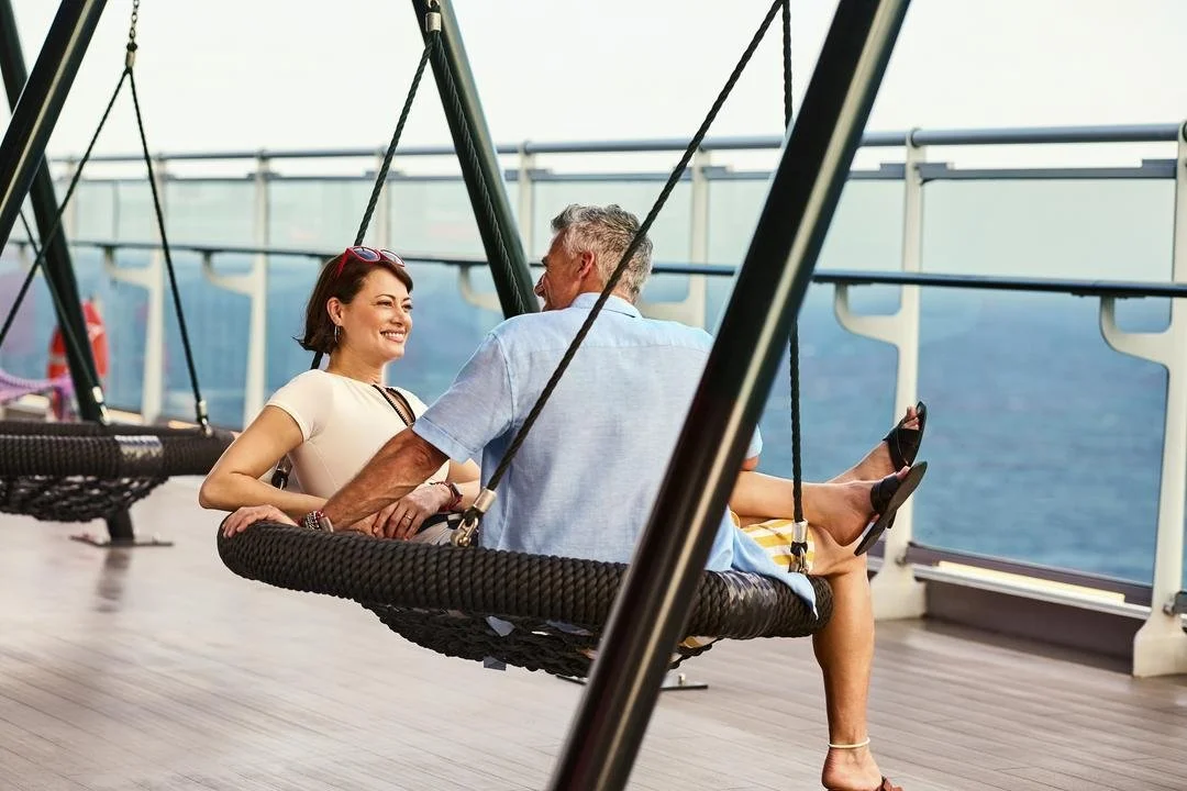 A woman and a man sitting on a swing on a cruise ship deck, smiling and talking, with ocean in the background.