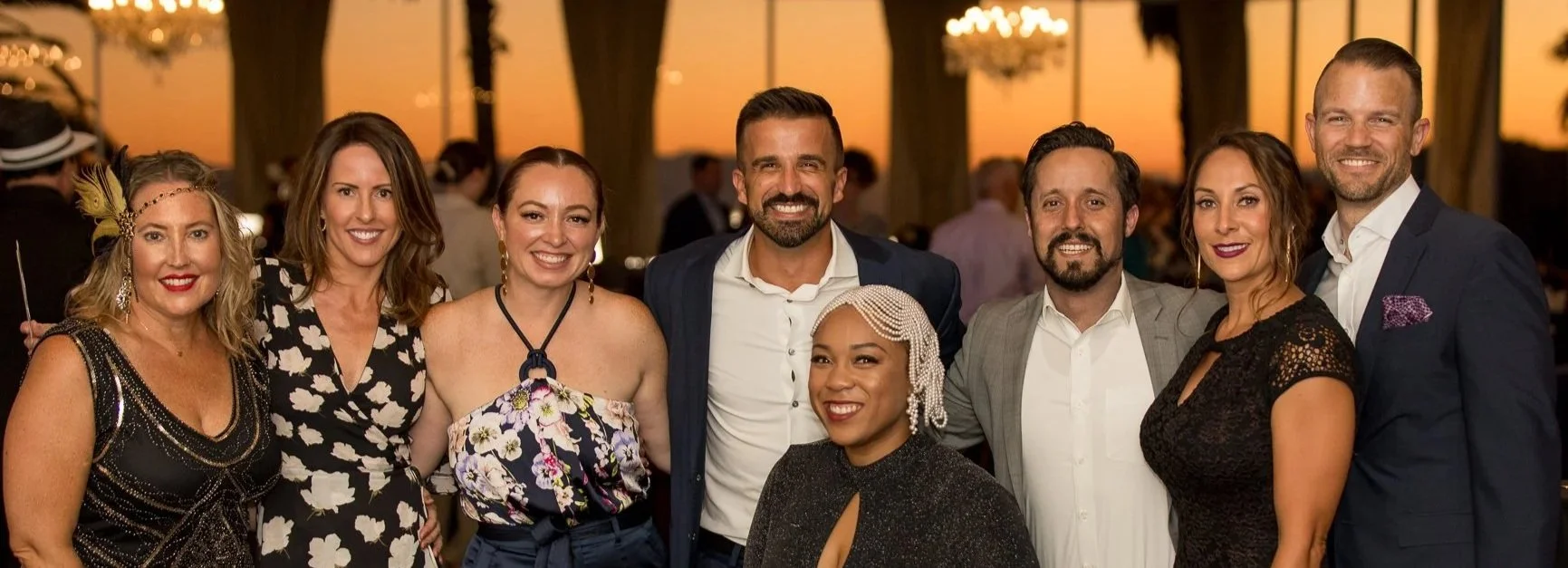 Group of nine people dressed in formal attire at an outdoor event during sunset, smiling for a photo.