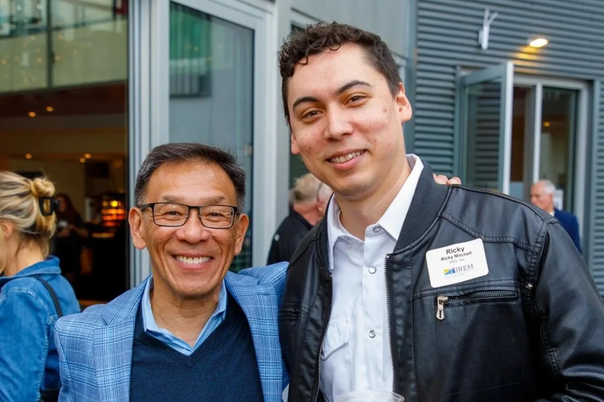 Two men smiling at an event, one wearing glasses and a blue blazer, the other wearing a black leather jacket and a name tag that reads 'Ricky'.