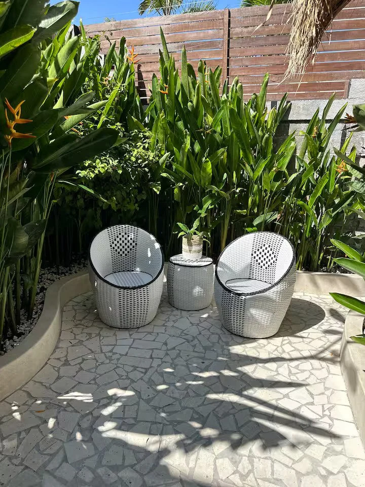 Two white circular outdoor chairs with black trim and a small matching side table with a potted plant on top, set on a stone-tiled patio surrounded by lush green plants and tall tropical foliage.