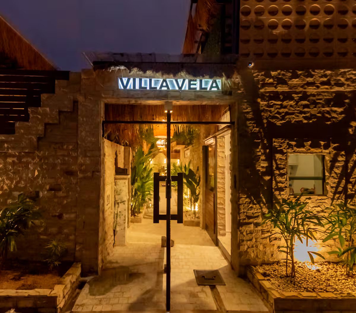 Night view of the entrance to a restaurant or bar with stone walls, illuminated sign that reads 'VICAVELA,' and warm lighting inside with potted plants along the walkway.