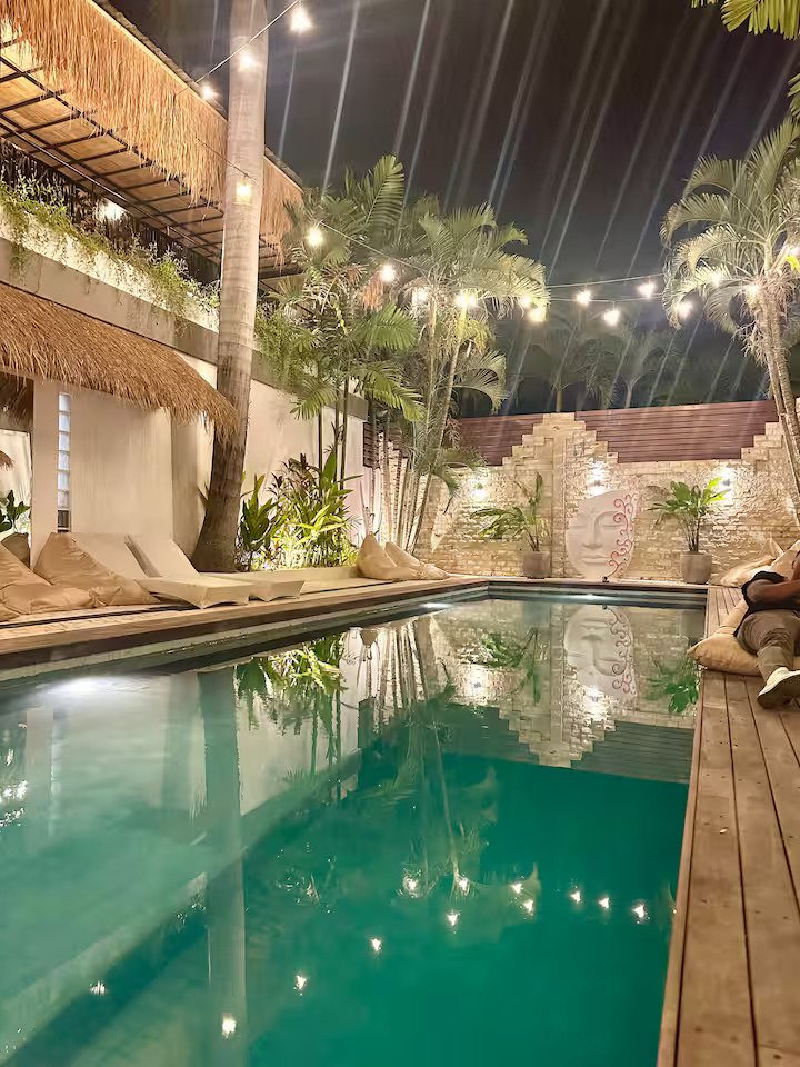 Nighttime view of a backyard pool area with lounge chairs, palm trees, string lights, and a decorative brick wall.