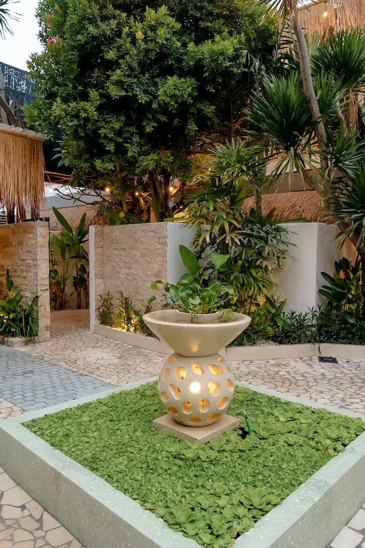 A decorative outdoor fountain with a plant on top, surrounded by green ground cover, in a lush garden with various plants and trees, enclosed by a low wall and stone pathway.