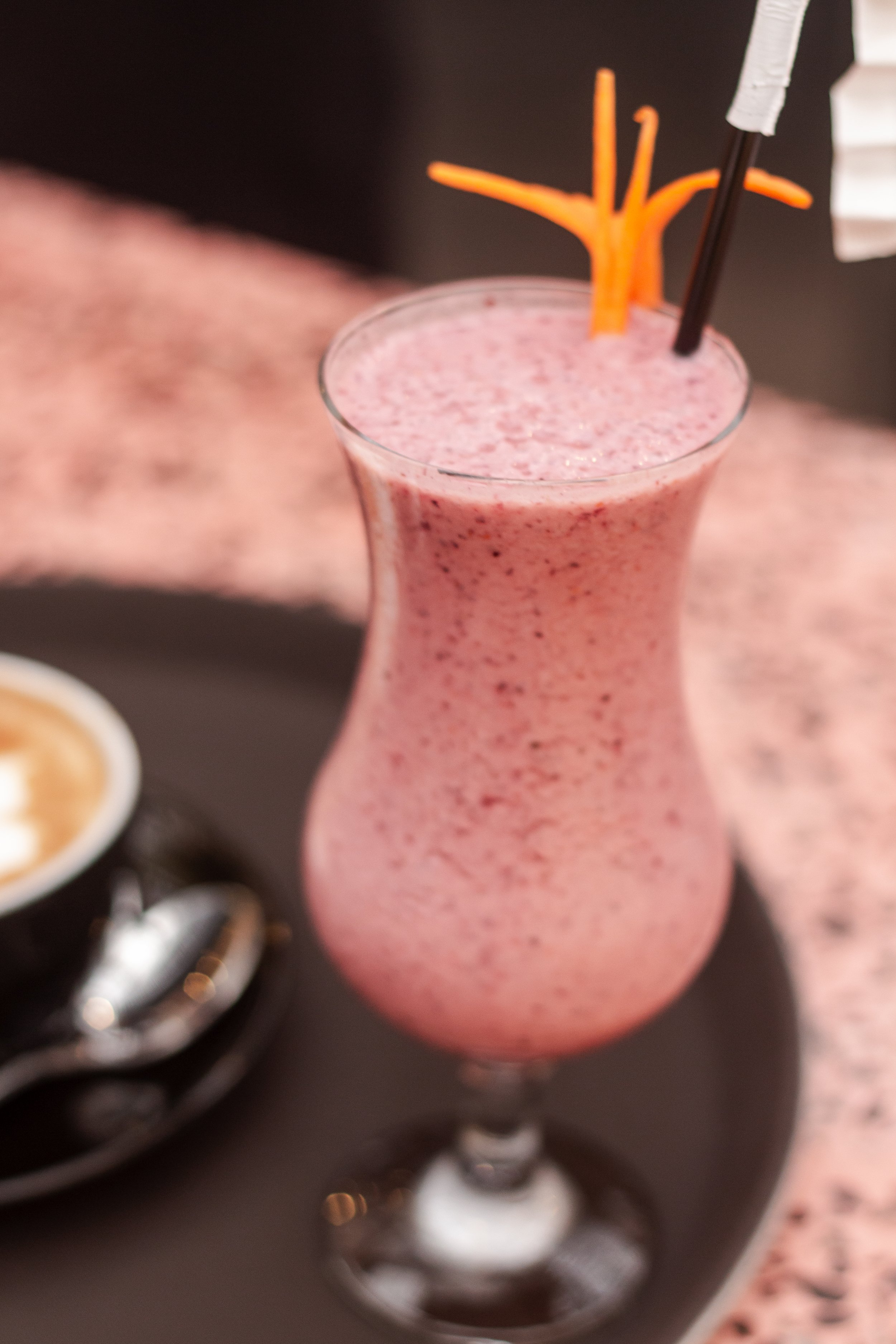 A pink, frothy smoothie in a tall glass with a decorative orange straw, placed on a black tray, accompanied by a small cup of coffee and a spoon.