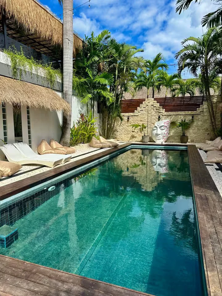 A backyard with a rectangular swimming pool surrounded by a wooden deck, lounge chairs with cushions, lush green plants, palm trees, and a decorative face sculpture on a stone wall.