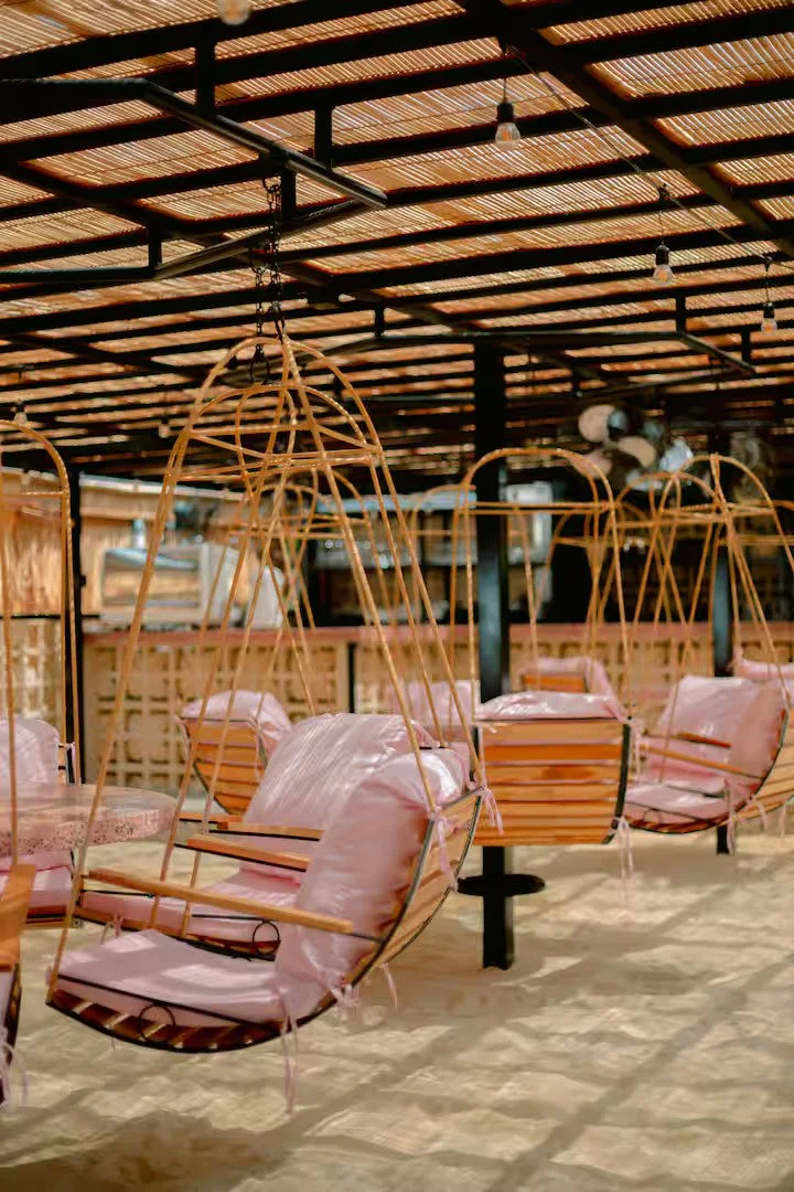 Outdoor seating area with hanging wicker chairs and cushions, sand floor, wooden and metal structure with a slatted roof.