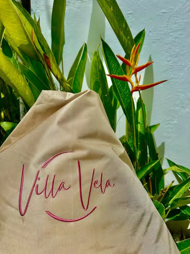 Close-up of a beige tote bag with pink embroidered text that reads 'Villa Vela,' surrounded by green tropical plants with long, pointed leaves and orange-red flowers.