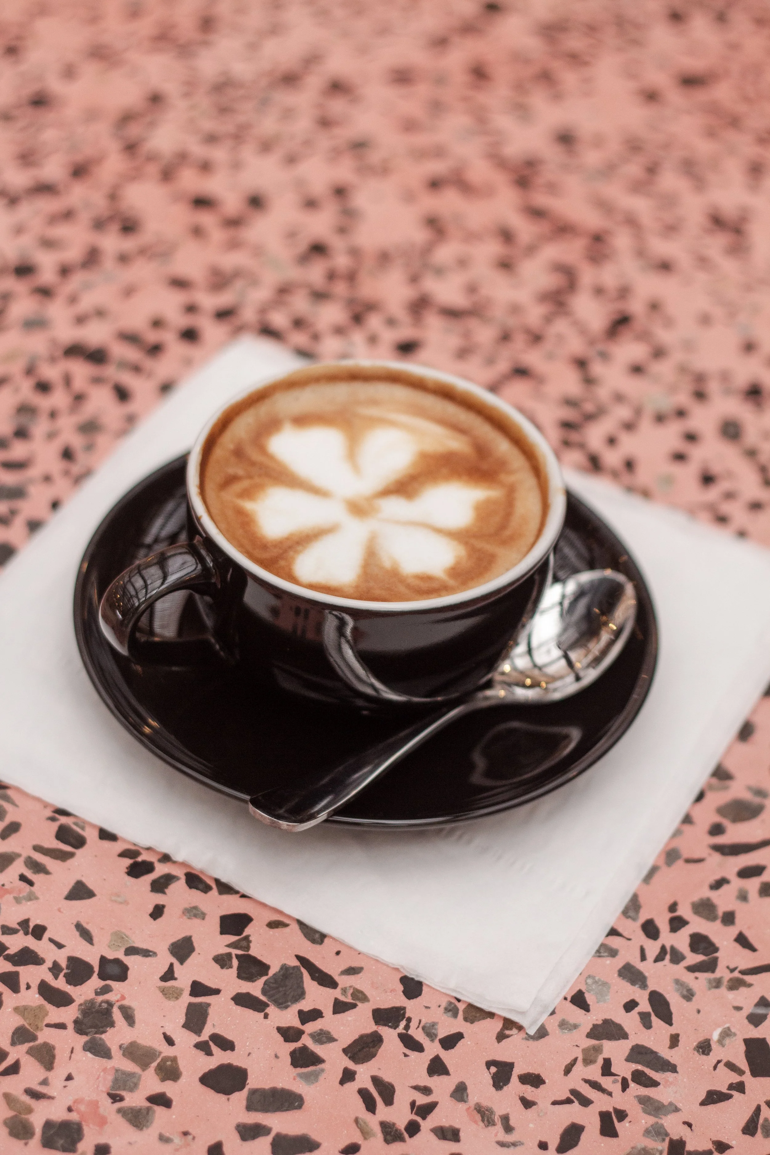 A black ceramic cup of cappuccino with latte art in the shape of a flower, sitting on a matching black saucer with a silver spoon on a white napkin on a pink and black terrazzo table.