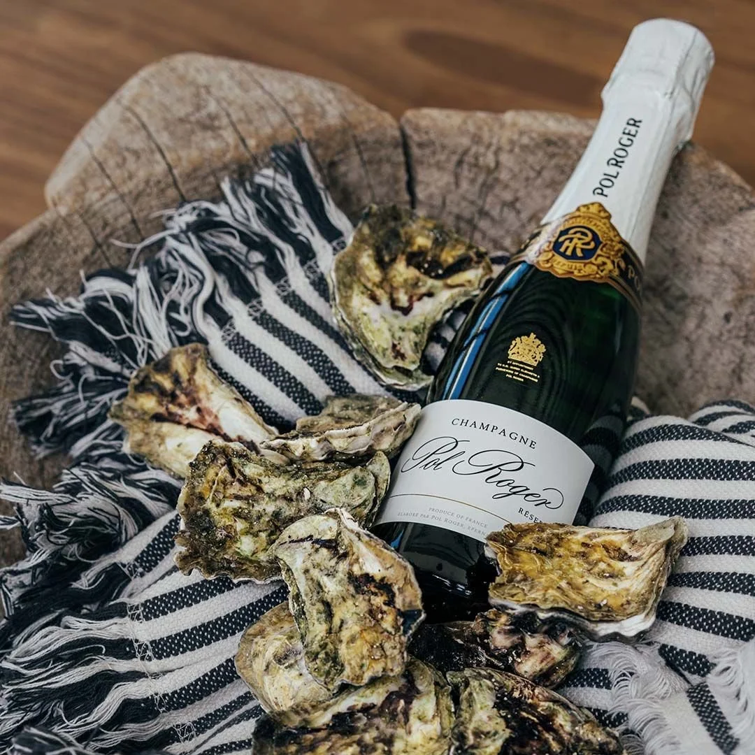 A bottle of Pol Roger champagne surrounded by oysters on a striped cloth, placed on a wooden surface.