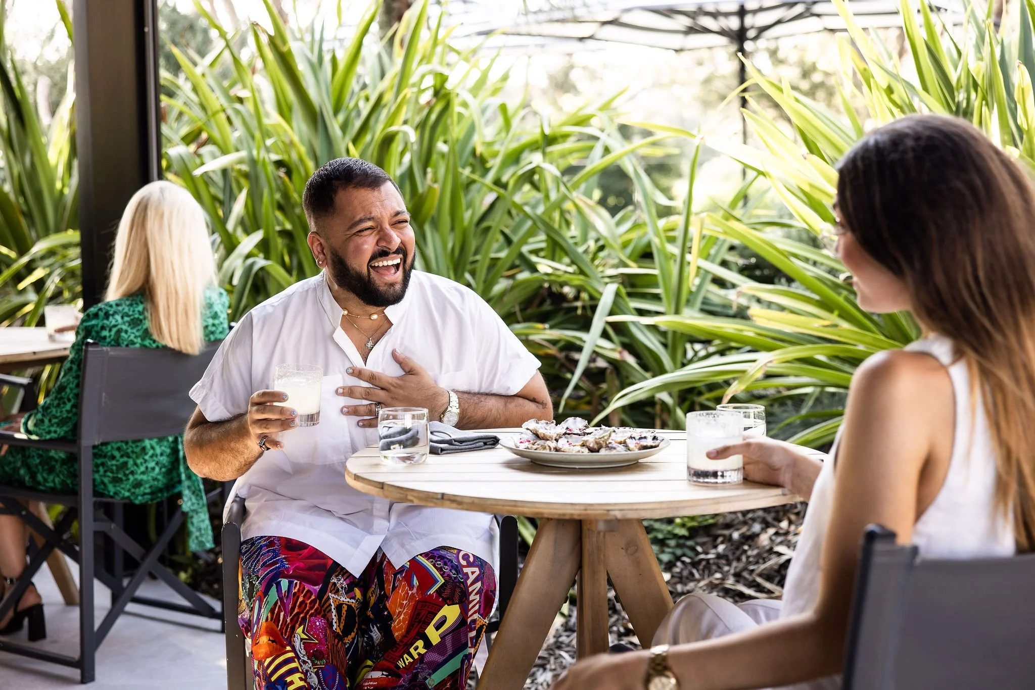 Two people are sitting at a round wooden table outdoors, smiling and enjoying drinks. The man is holding a glass of milk and has his hand on his chest, appearing to laugh or share a joyful moment. The woman is facing him, holding a glass and smiling. There is a plate of food on the table, and lush green plants surround them. In the background, another woman with blonde hair and a green patterned dress is sitting at a different table.