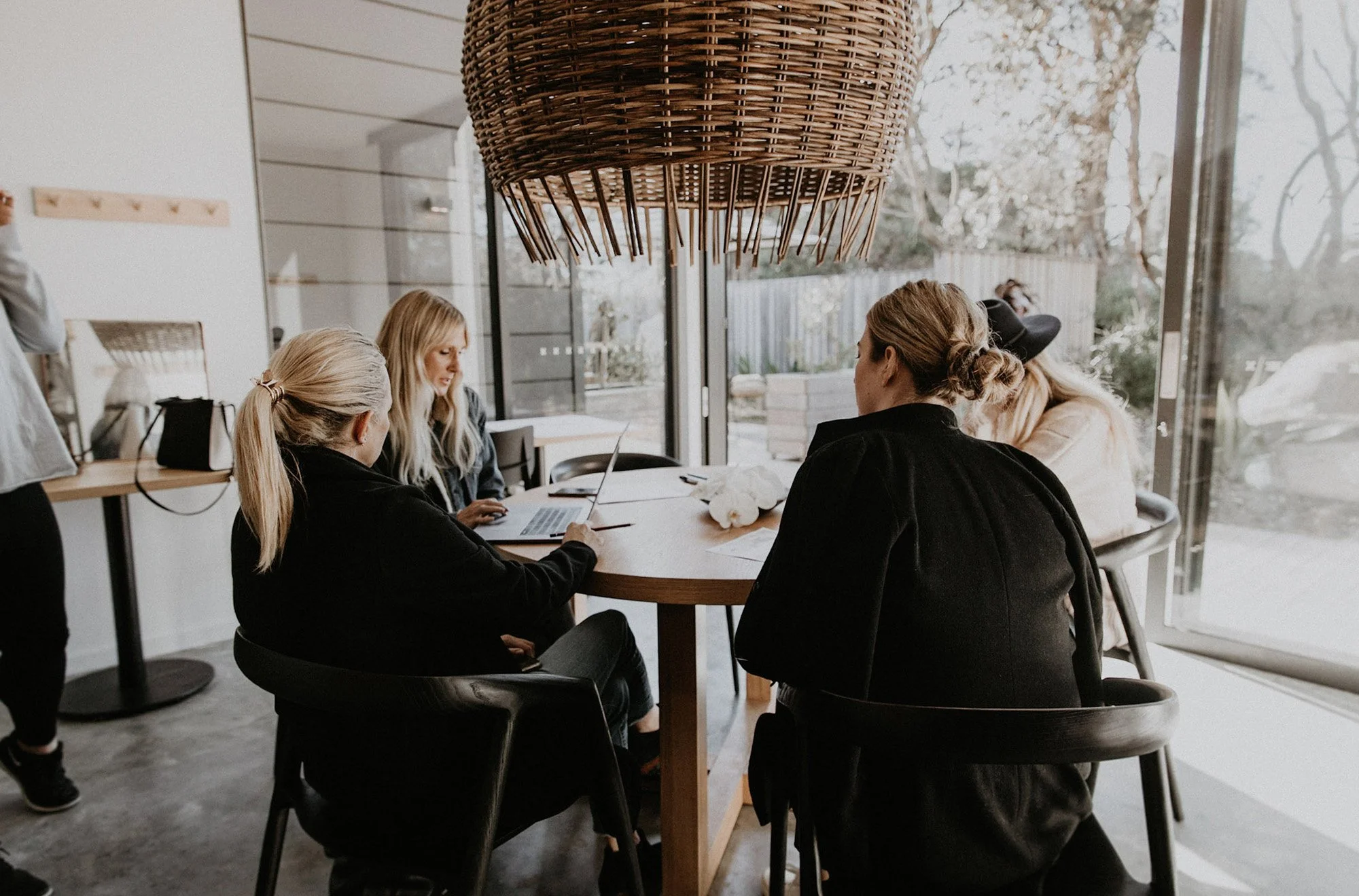 Four women sitting around a wooden table in a modern, well-lit room with large glass doors showing outdoor trees and sky.