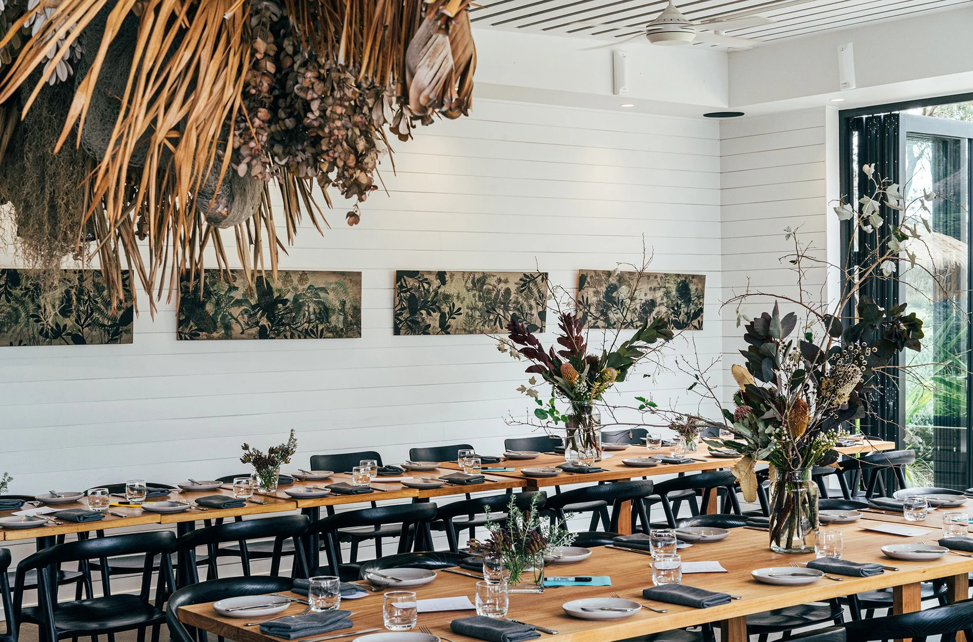 A modern dining room set up for a gathering with long wooden tables, black chairs, and large floral arrangements in glass vases. The table settings include white plates, black napkins, and glasses, with artwork on the white paneled walls and large wi