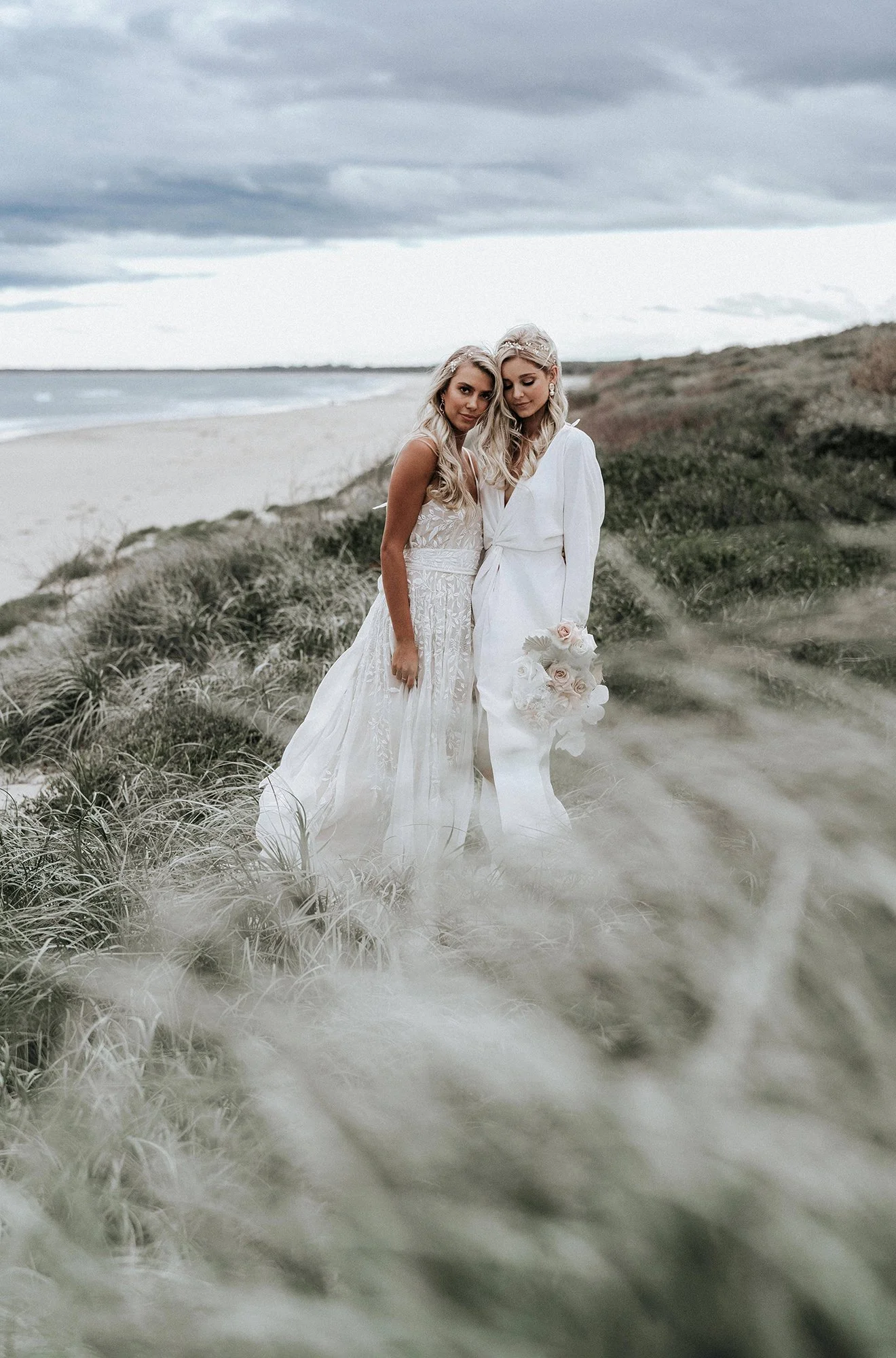 Two women in white dresses standing on a beach with grassy dunes and cloudy sky in the background, one holding a bouquet of pale pink roses.