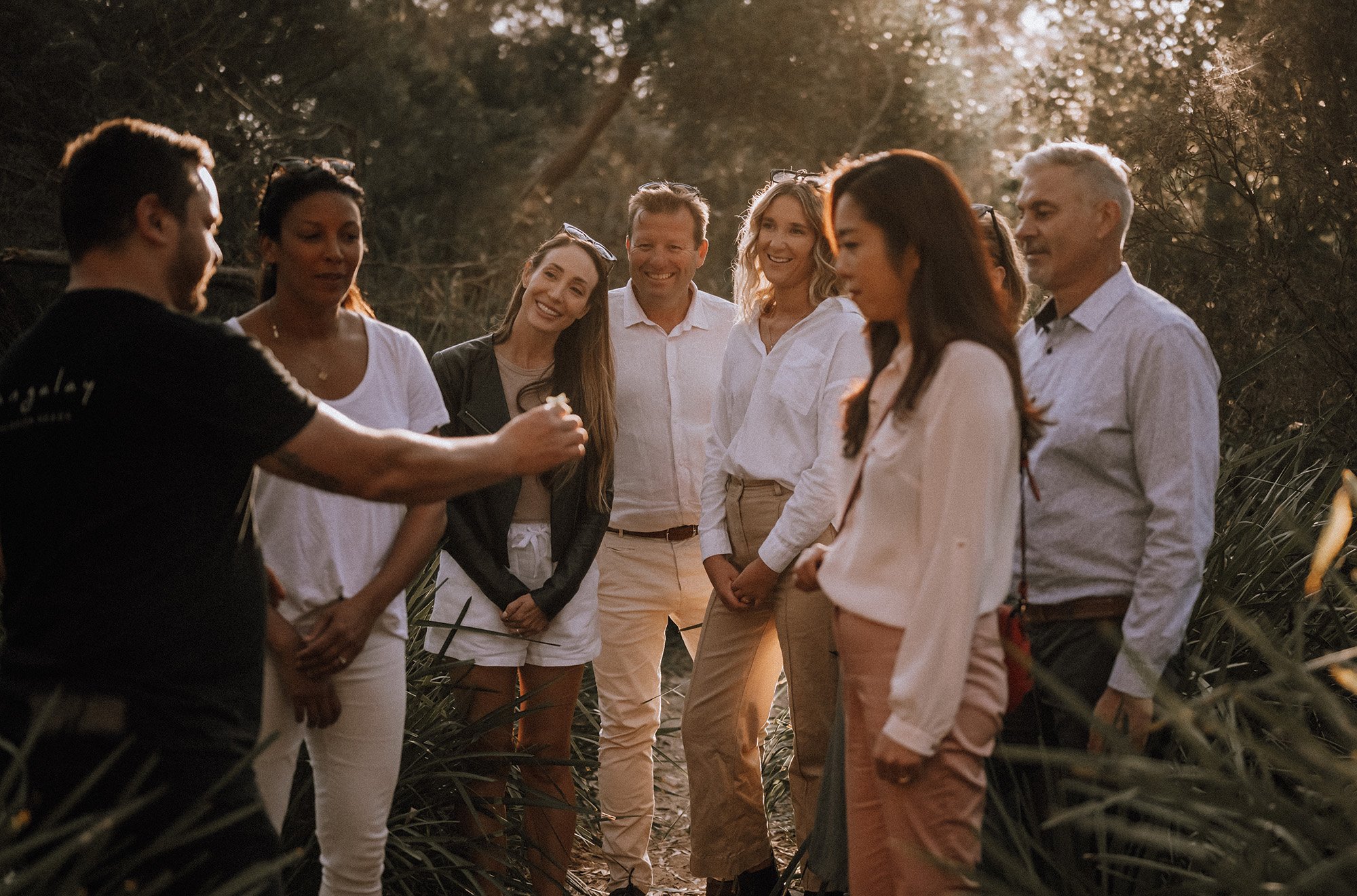 A group of people standing outdoors in a field, listening to a man speak with a microphone, during sunset or late afternoon. They are smiling and engaged.