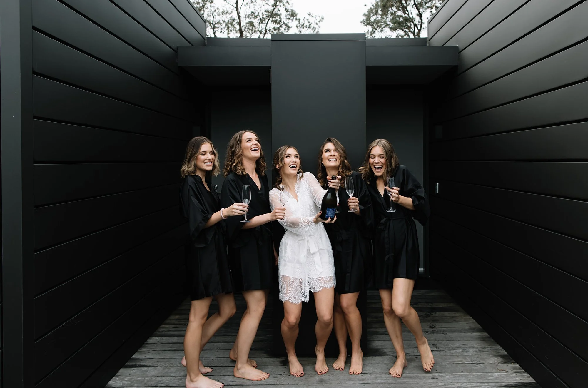 Five women in black robes, with one in a white lace robe, celebrating with champagne in an outdoor black-walled area.