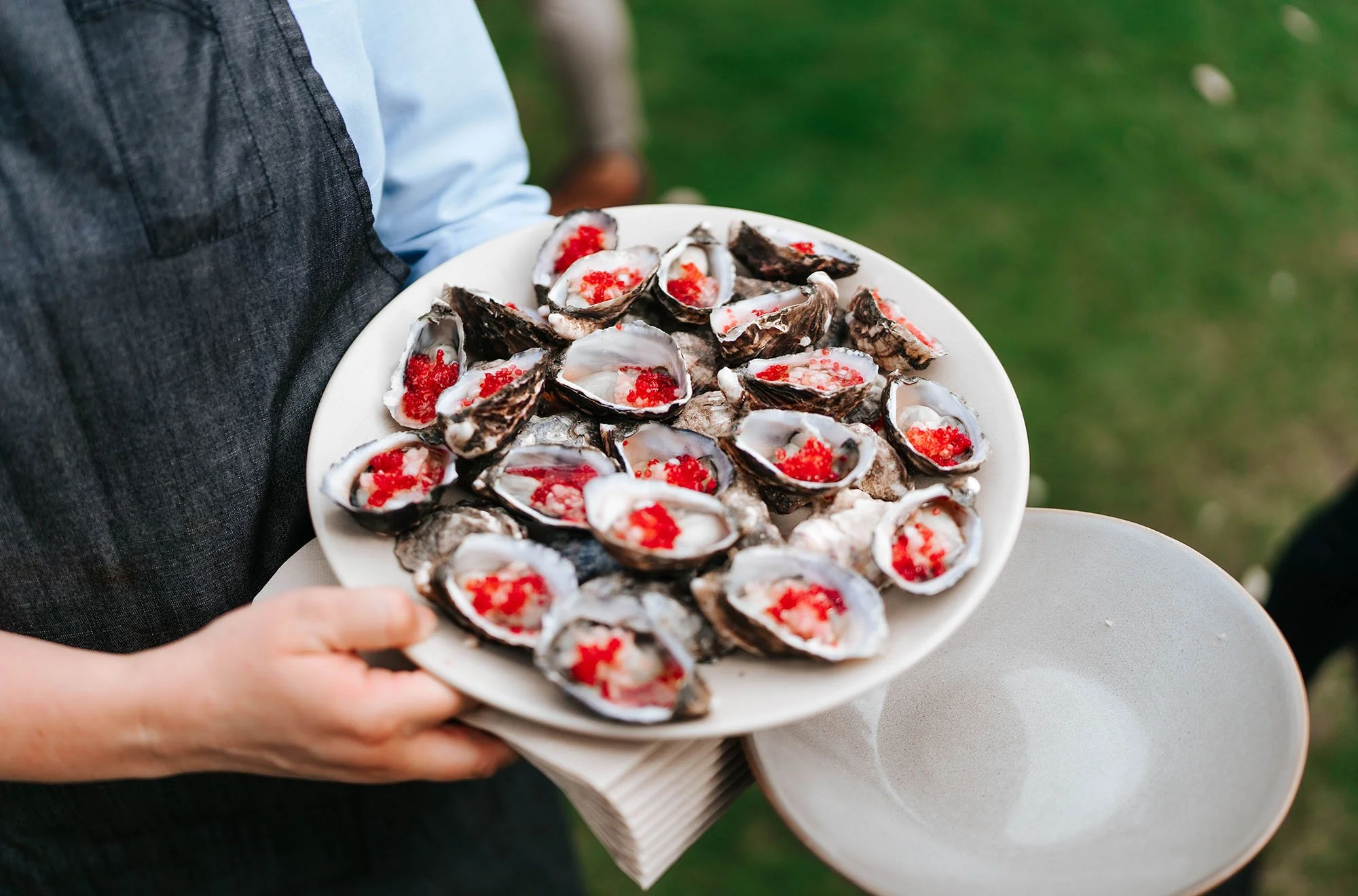 Person holding a plate of opened oysters topped with red fish roe, with an empty side plate in the foreground, outdoors on grass.