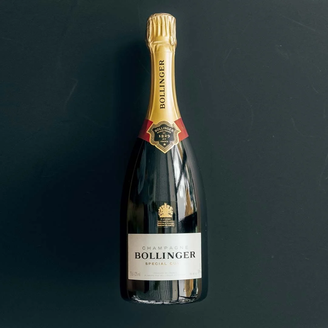 A bottle of Bollinger champagne with a gold foil cap, a black and gold neck label, and a white front label, placed on a black surface.
