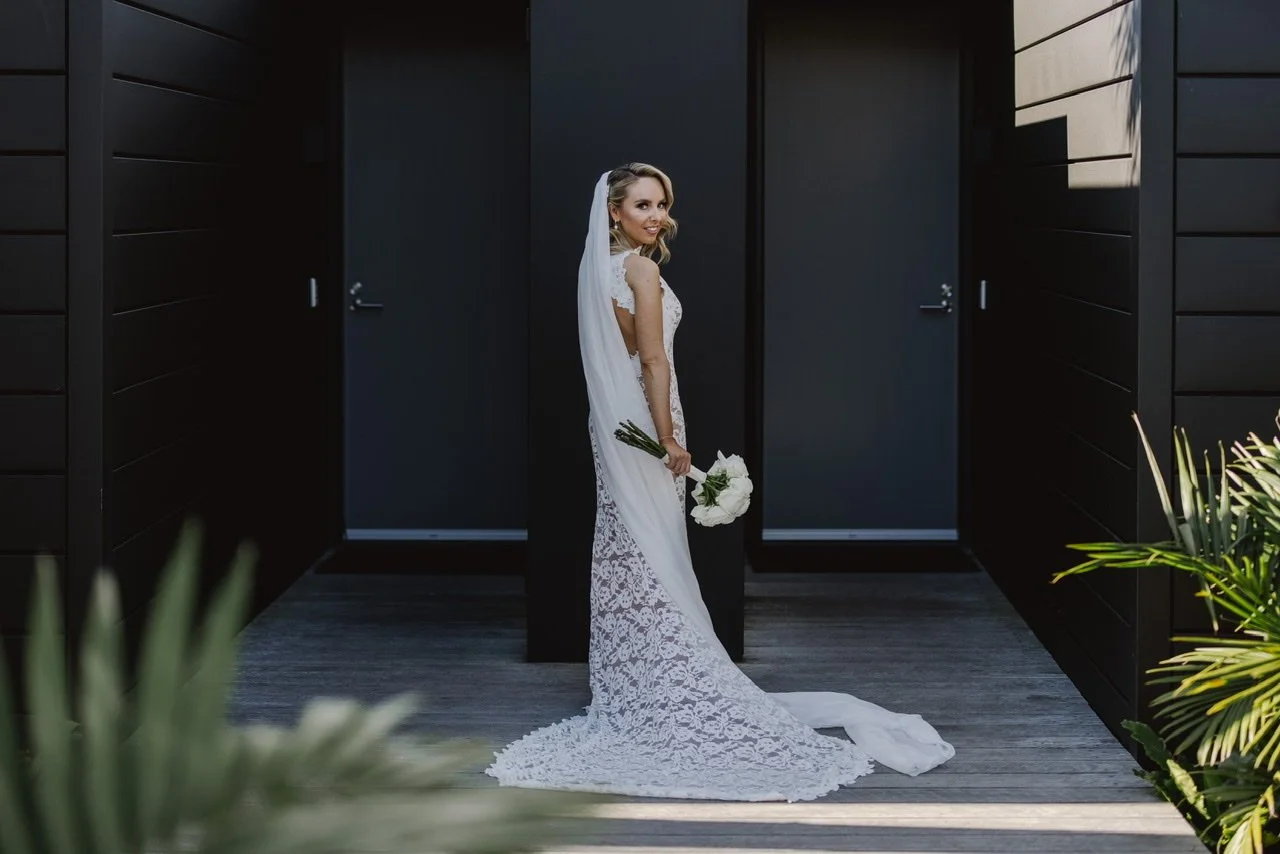 Bride in a white lace wedding gown holding a bouquet of white flowers standing between black doors with green plants in the foreground.