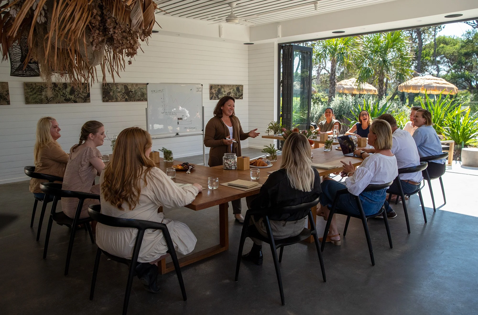 A woman standing and speaking to a group of people seated at a conference table in a bright room with large windows showing outdoor greenery.