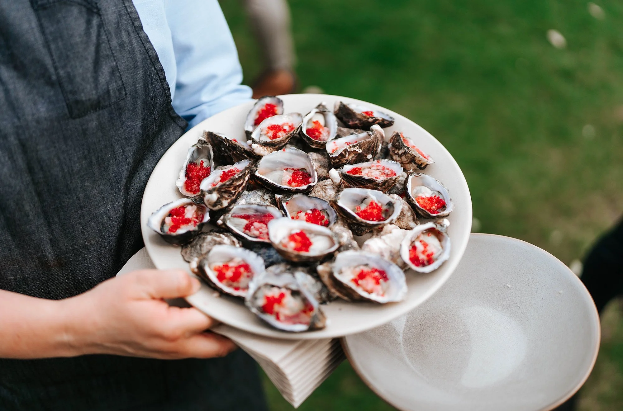 Person holding a plate of oysters topped with red fish roe, with an empty plate underneath, outdoors on grass.