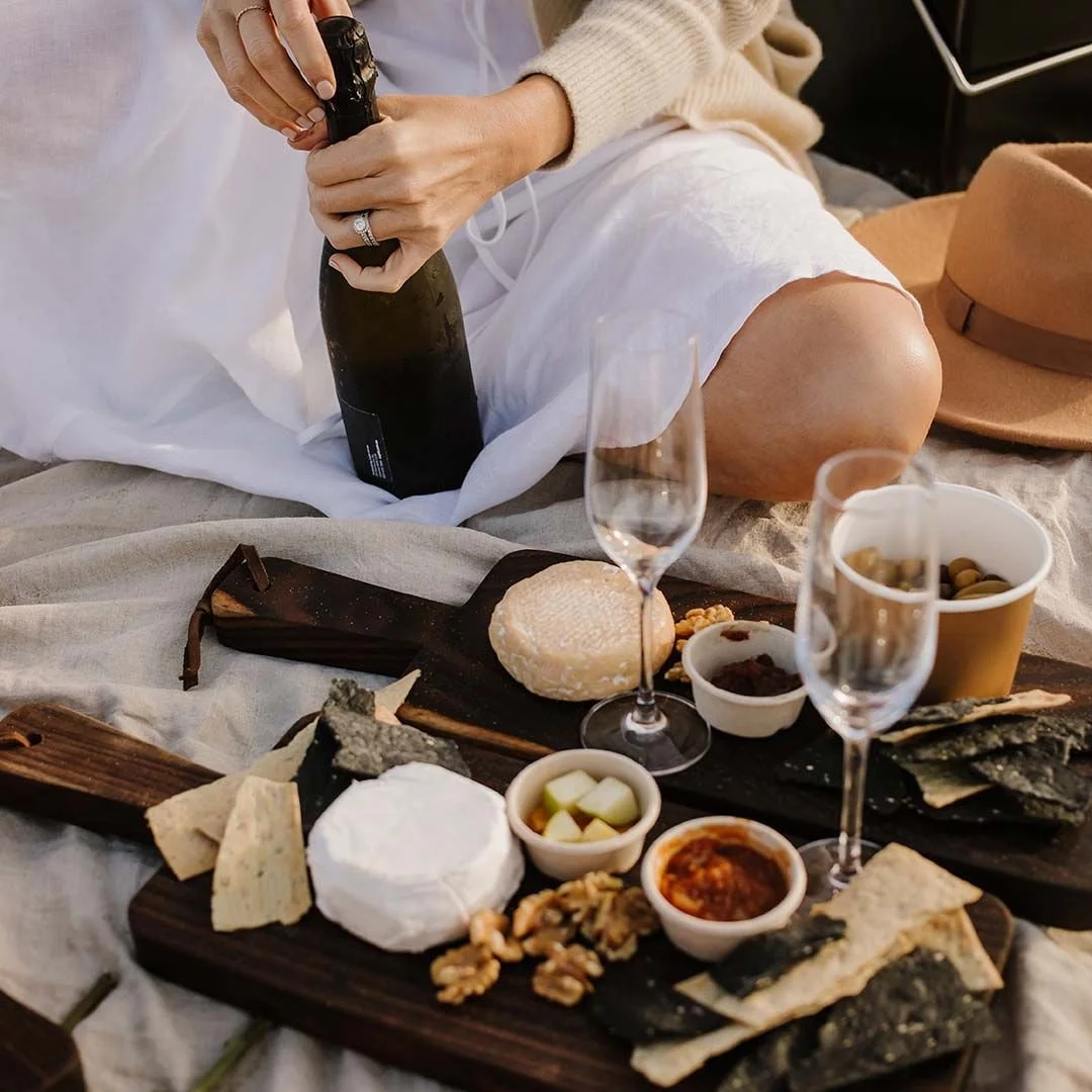 A woman sitting on a blanket with a cheese platter, champagne, and snacks.