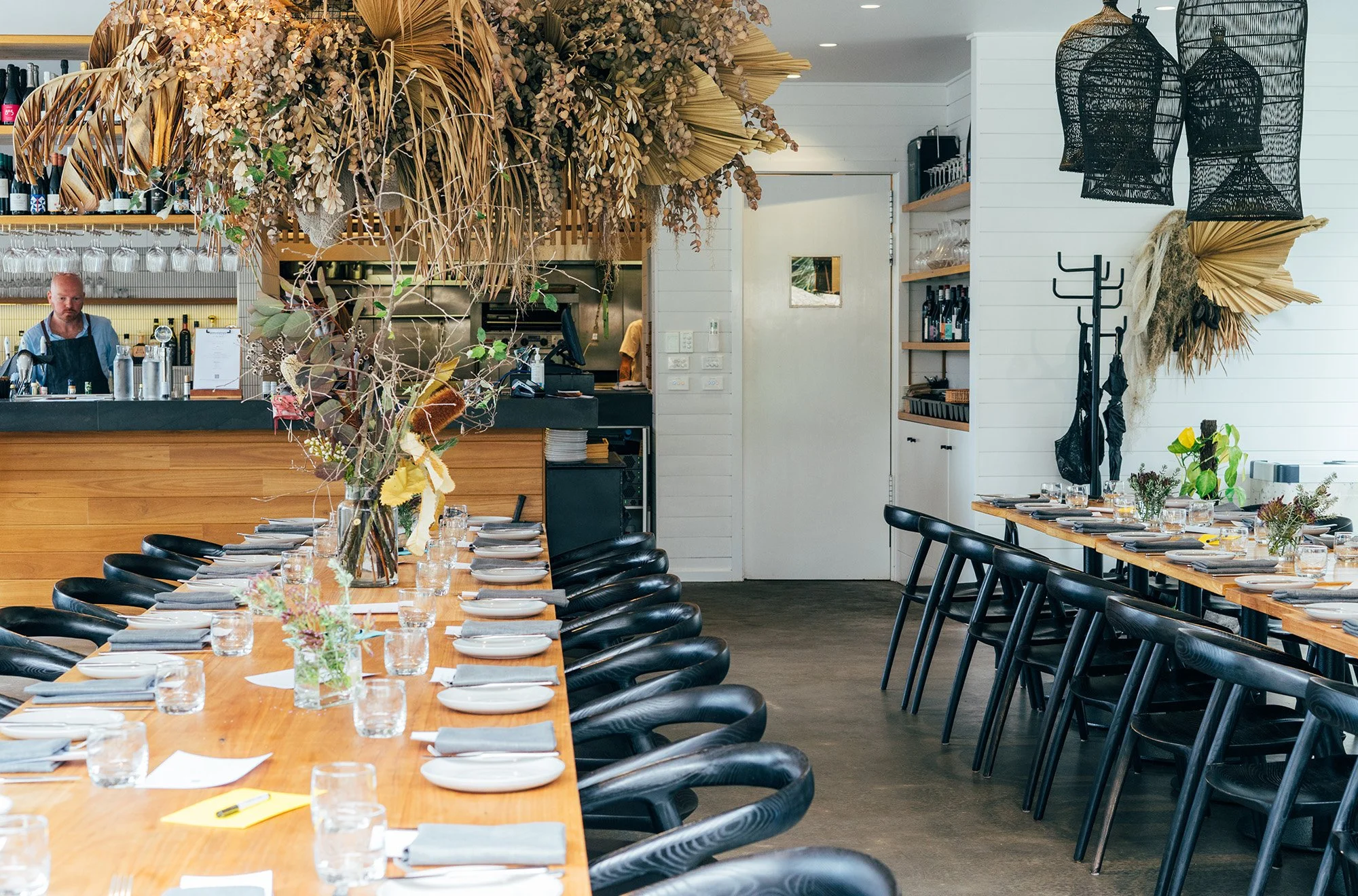 Elegant restaurant interior with a long wooden table set with plates, glasses, and napkins, decorated with flower arrangements, black chairs, and decorative hanging lamps, featuring a bar area and a floral ceiling installation.