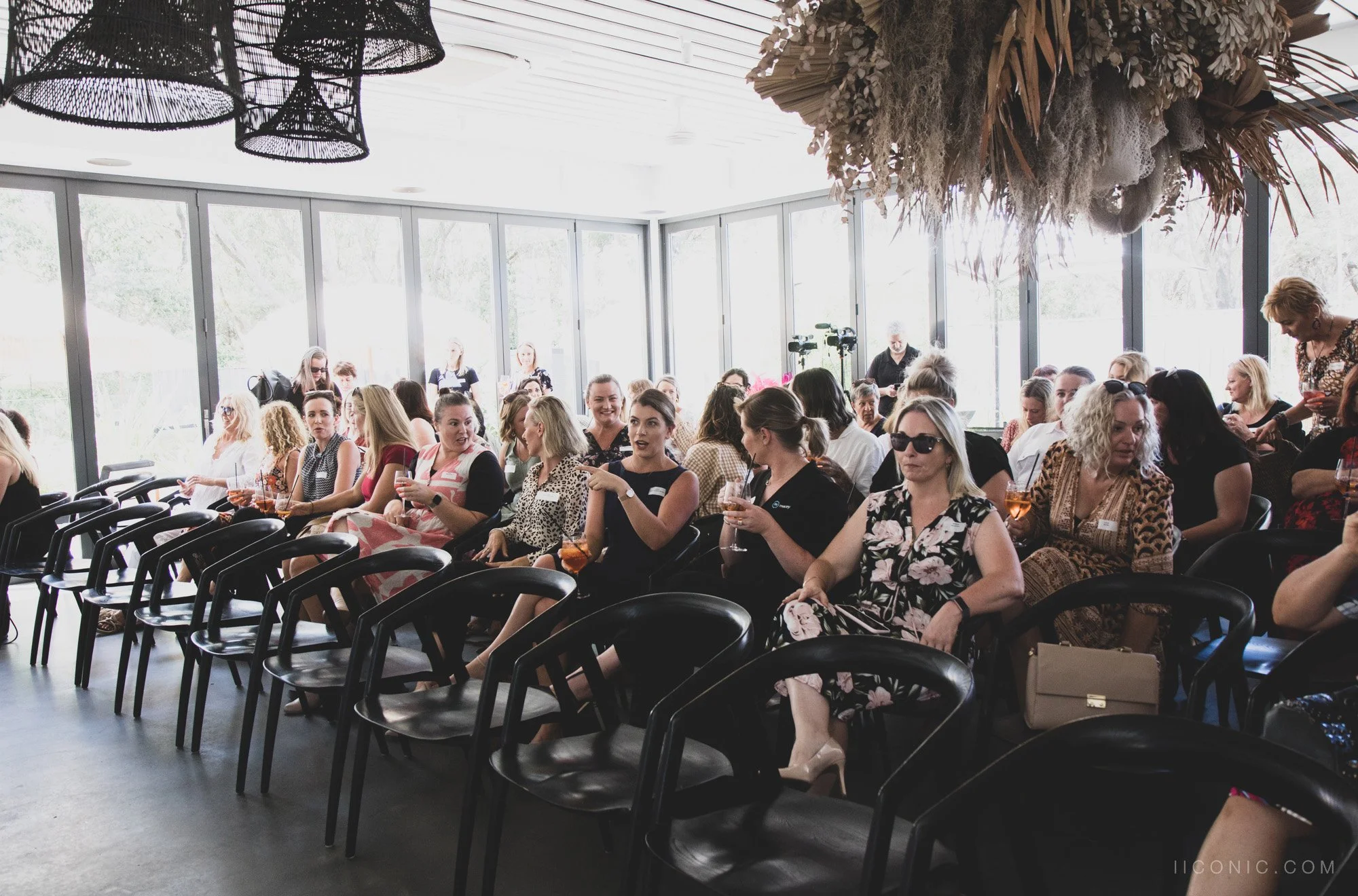 Women in a bright, modern conference room seated in black chairs, some holding glasses of wine, engaged in a discussion or listening to a speaker.