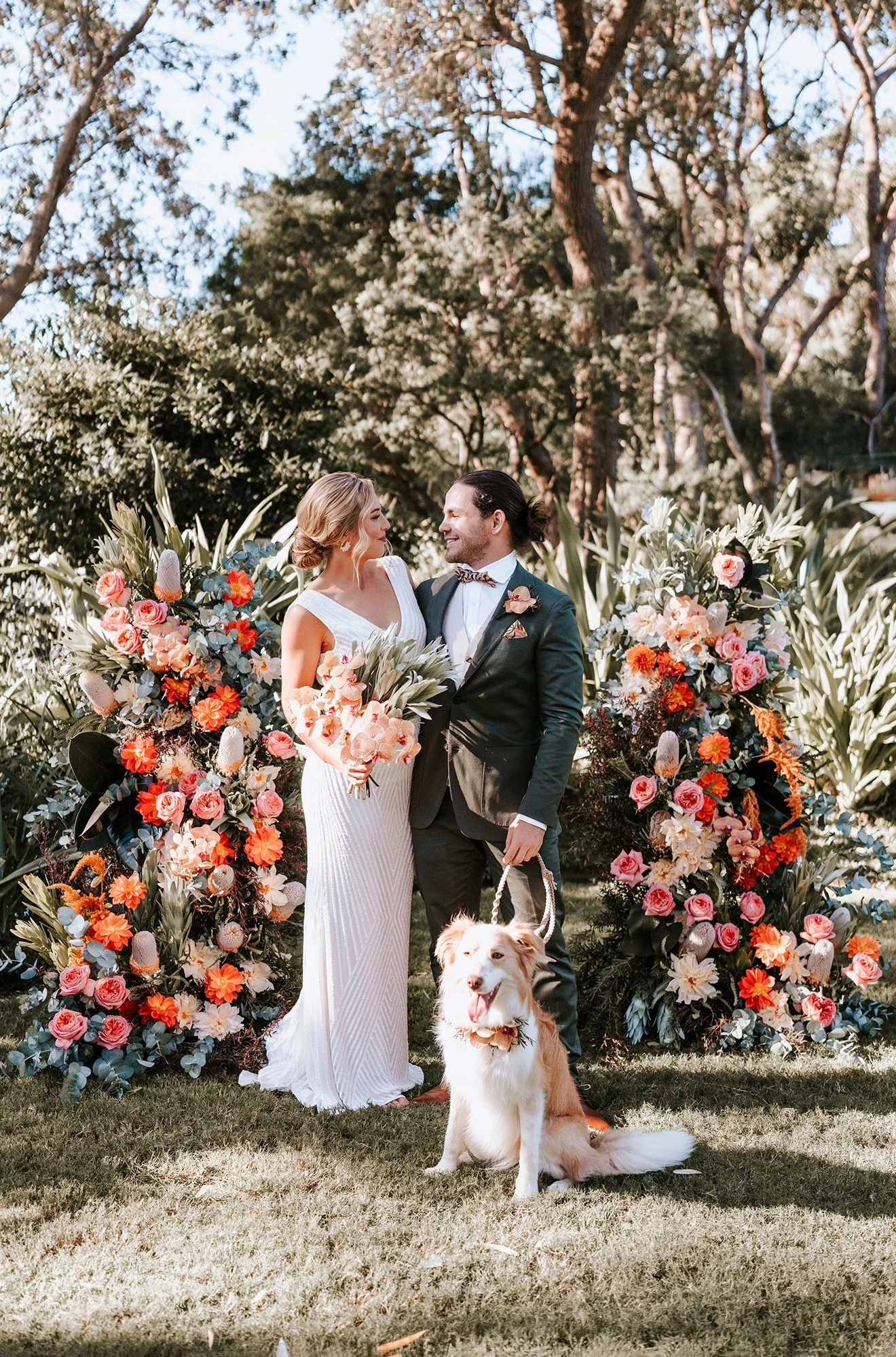 A bride and groom stand together outdoors surrounded by colorful floral arrangements, with a dog sitting in front of them. The bride wears a white dress, the groom in a dark suit with a bow tie, and they gaze at each other lovingly.