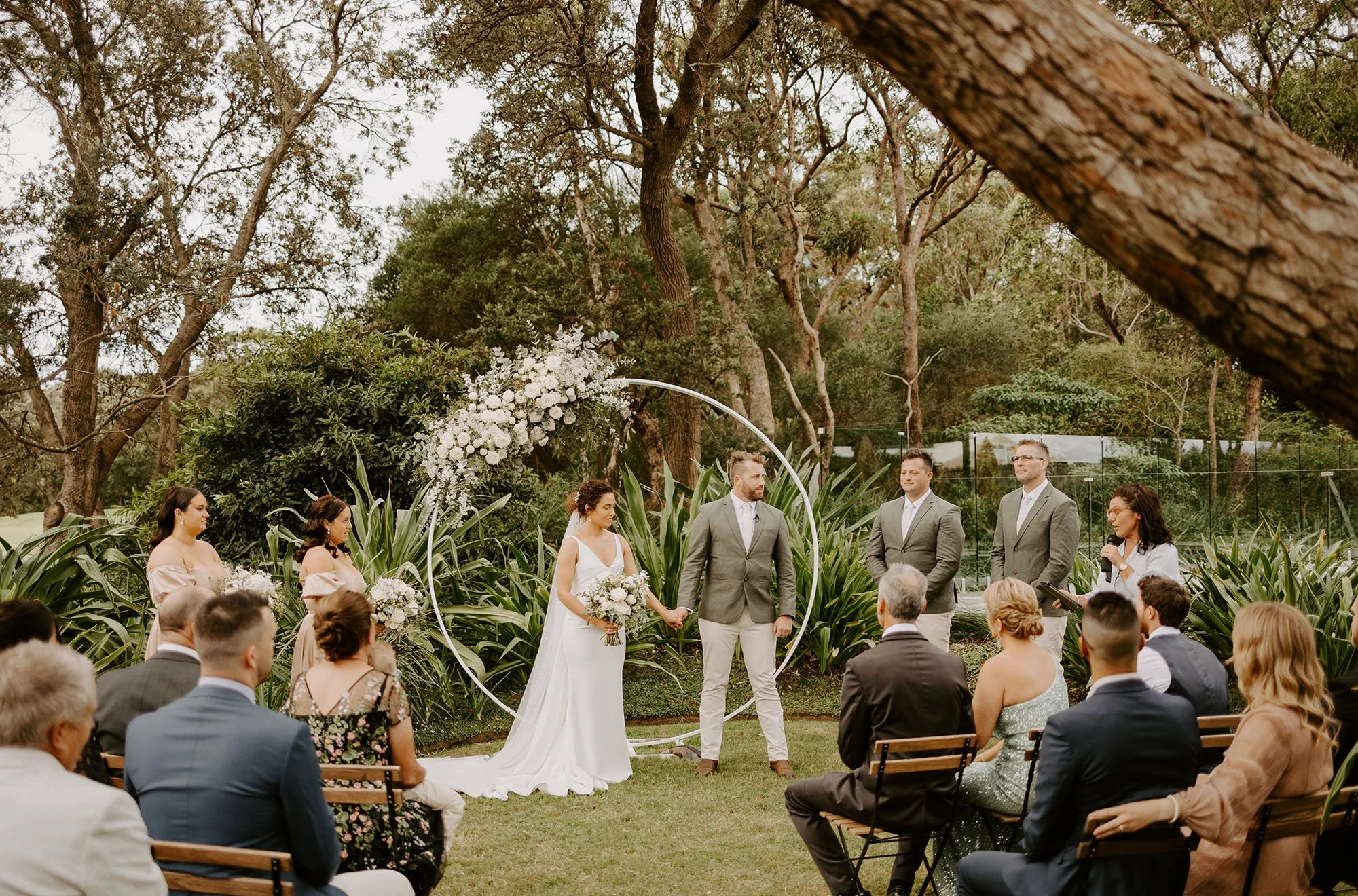 A wedding ceremony taking place outdoors with the bride and groom holding hands at the altar, surrounded by friends and family, and a floral arch in the background.