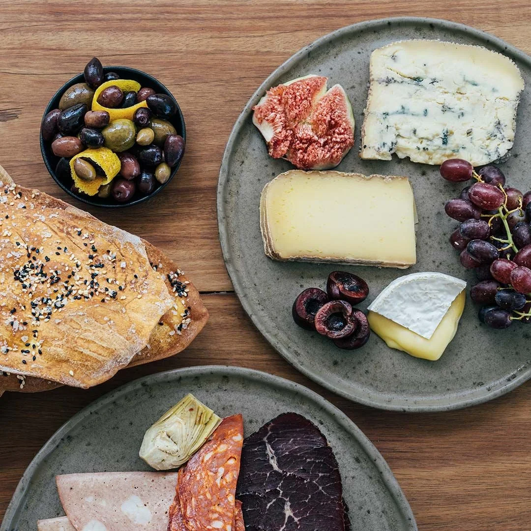 Cheese platter with various cheeses, grapes, and figs, alongside a loaf of seed-topped bread and a bowl of mixed olives, all arranged on rustic ceramic plates and a wooden table.