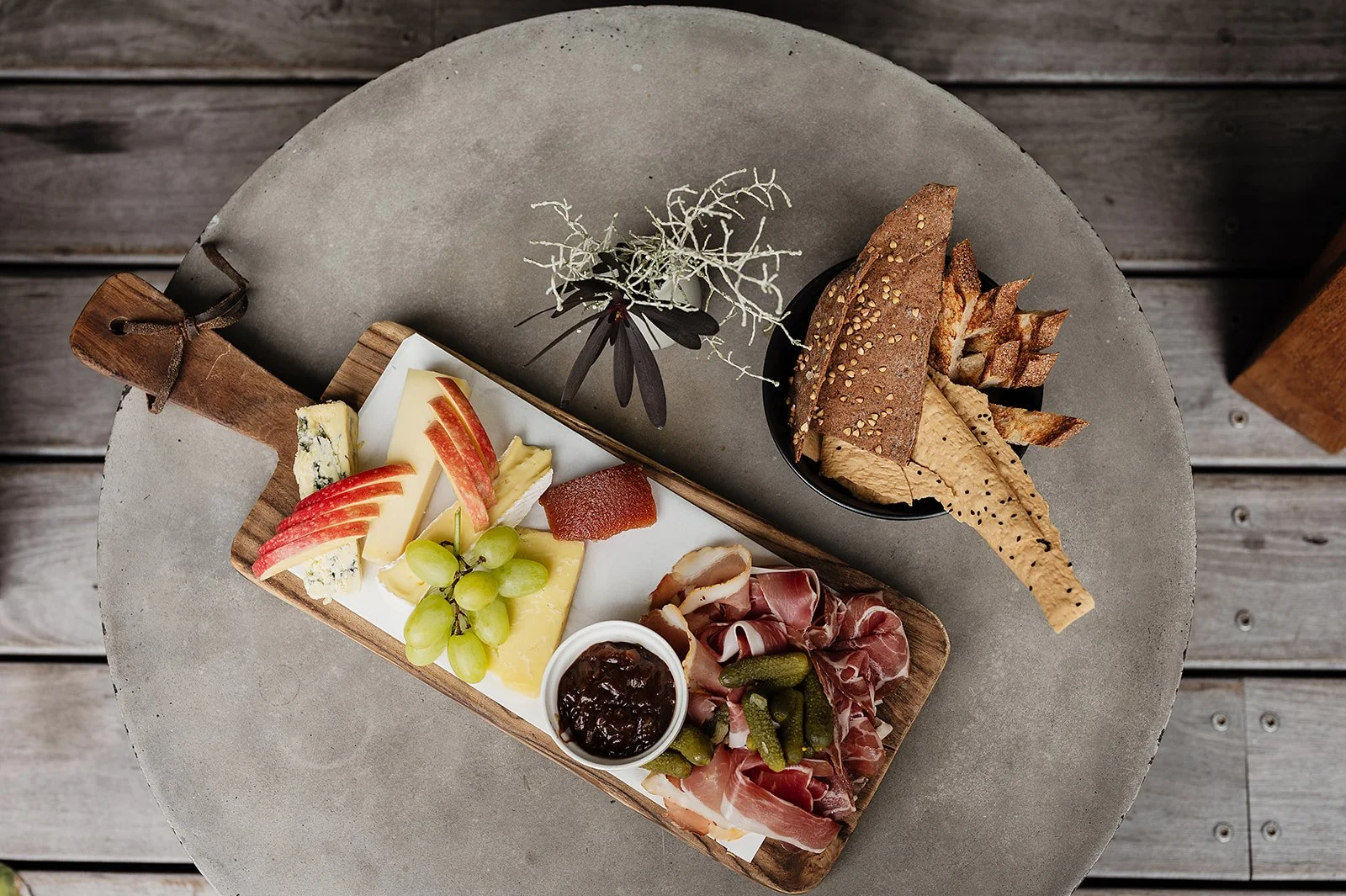Cheese platter with grapes and figs, a cheese with mold, a hard cheese, and a soft cheese, a bowl of olives, a loaf of sprinkled bread, and a meat platter with sliced cured meats and artichoke hearts on a wooden table.