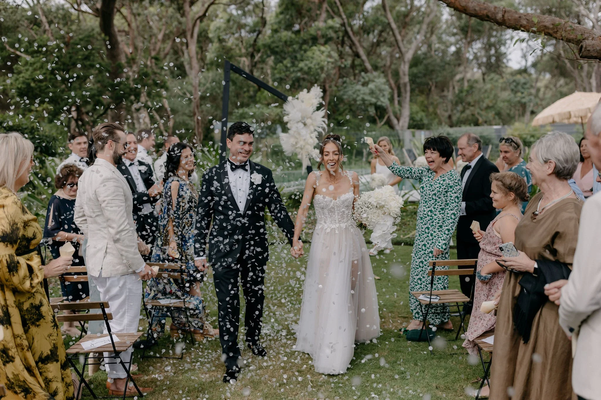 A newlywed couple walking hand in hand during their outdoor wedding celebration, surrounded by friends and family throwing confetti.