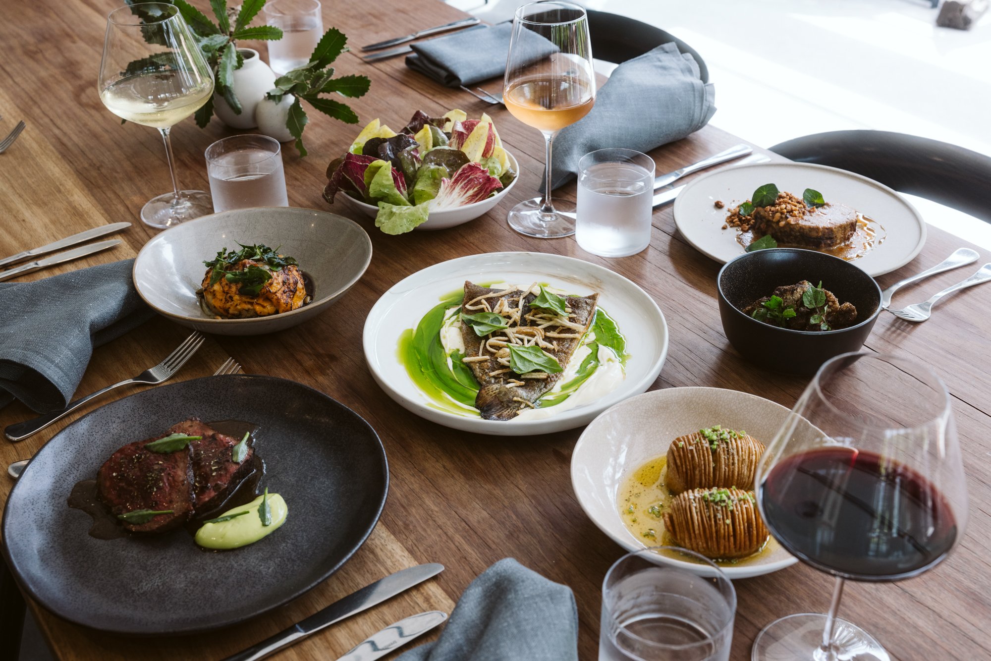 A table set with various dishes including a salad, a fish entree, a steak, a dessert, and glasses of wine and water in a sophisticated dining setting.