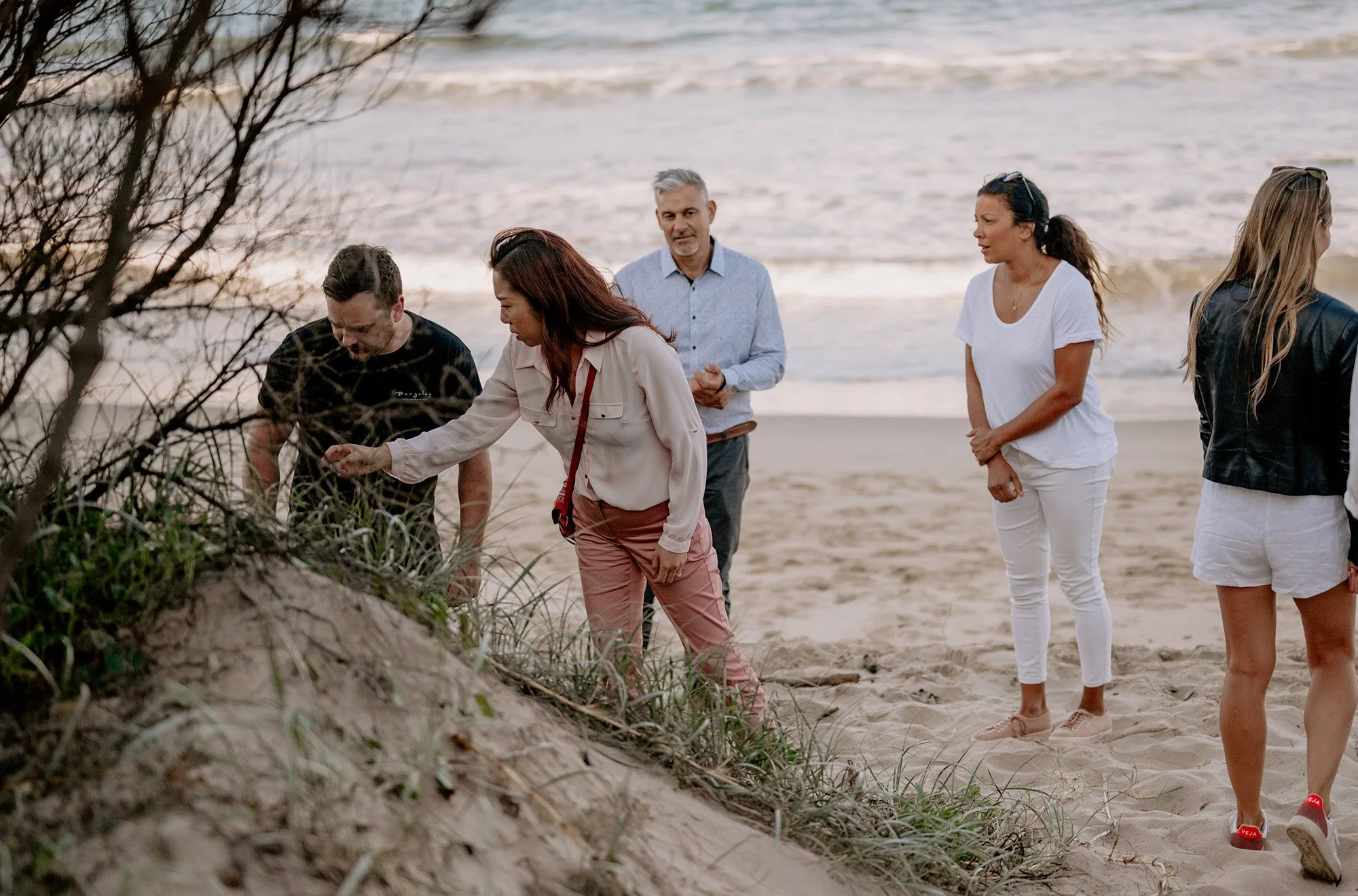 People walking along a sandy beach, some observing the sand dunes and plants in the foreground, with the ocean waves in the background.