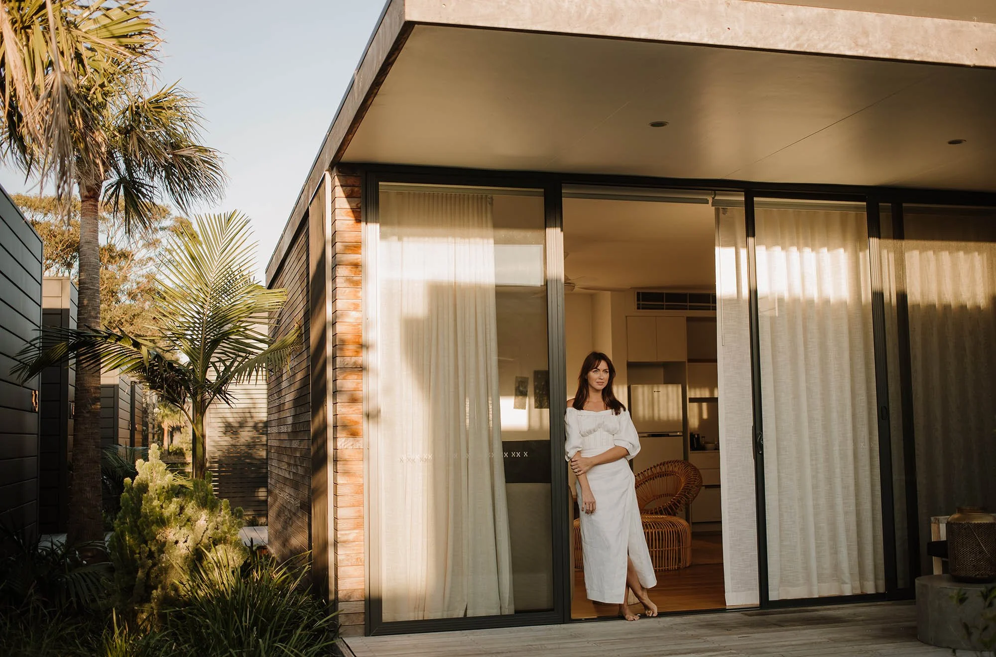 A woman standing at a large glass sliding door of a modern house, with sunlight streaming inside. The house has a wooden and brick exterior, surrounded by tropical plants and palm trees.