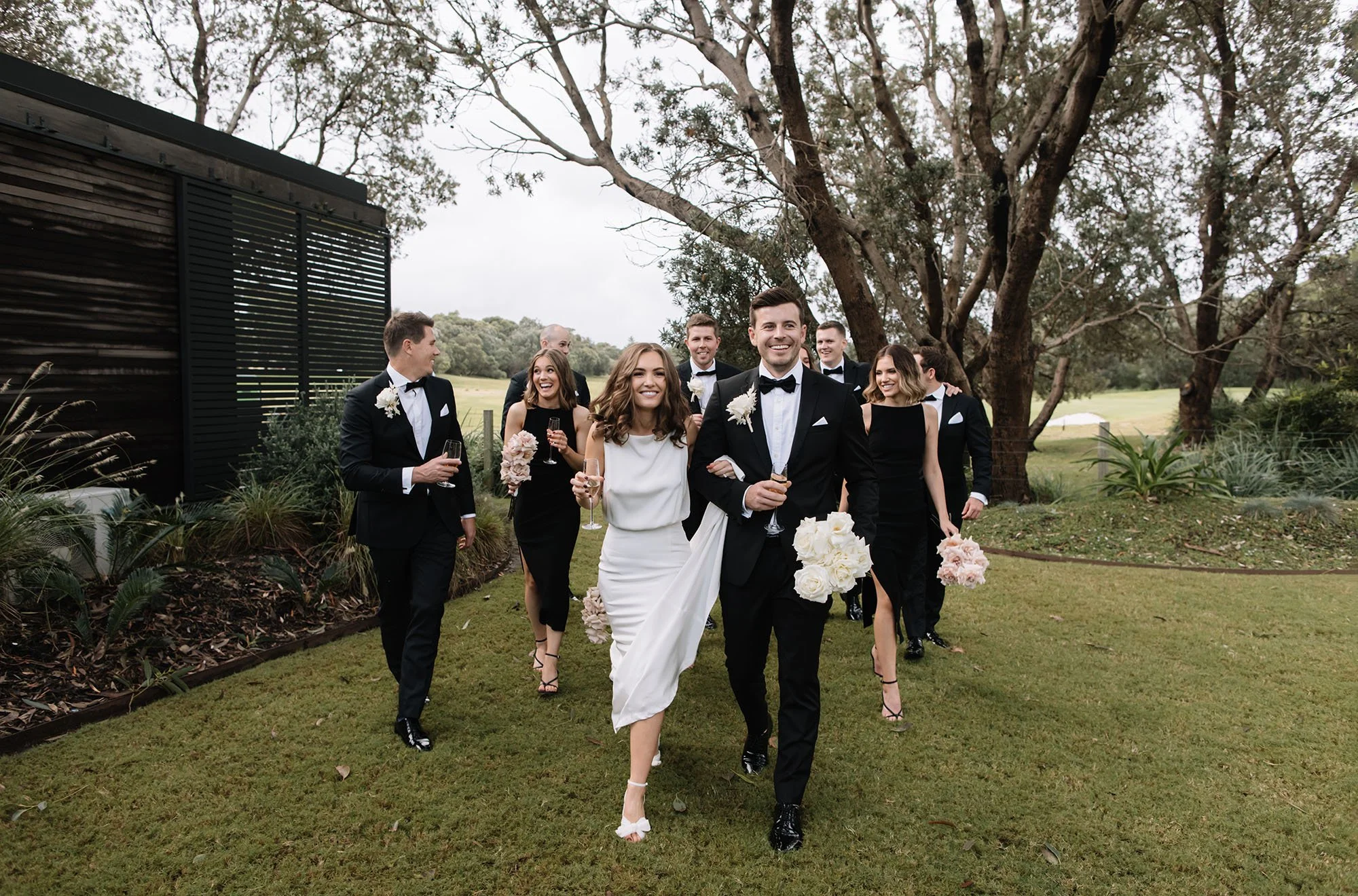 A wedding procession with a bride in a white dress and groom in a tuxedo leading the group, holding champagne glasses and flowers in an outdoor setting with trees and grass.