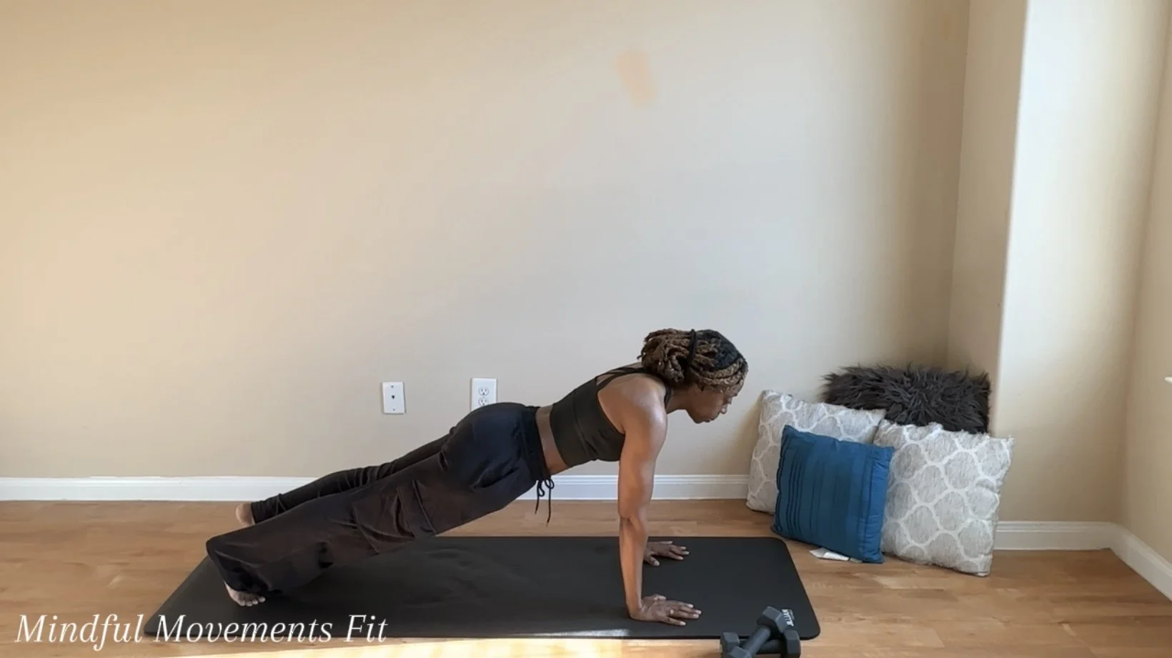 Woman performing a plank exercise on a yoga mat in a room with hardwood floors and pillows stacked against the wall.