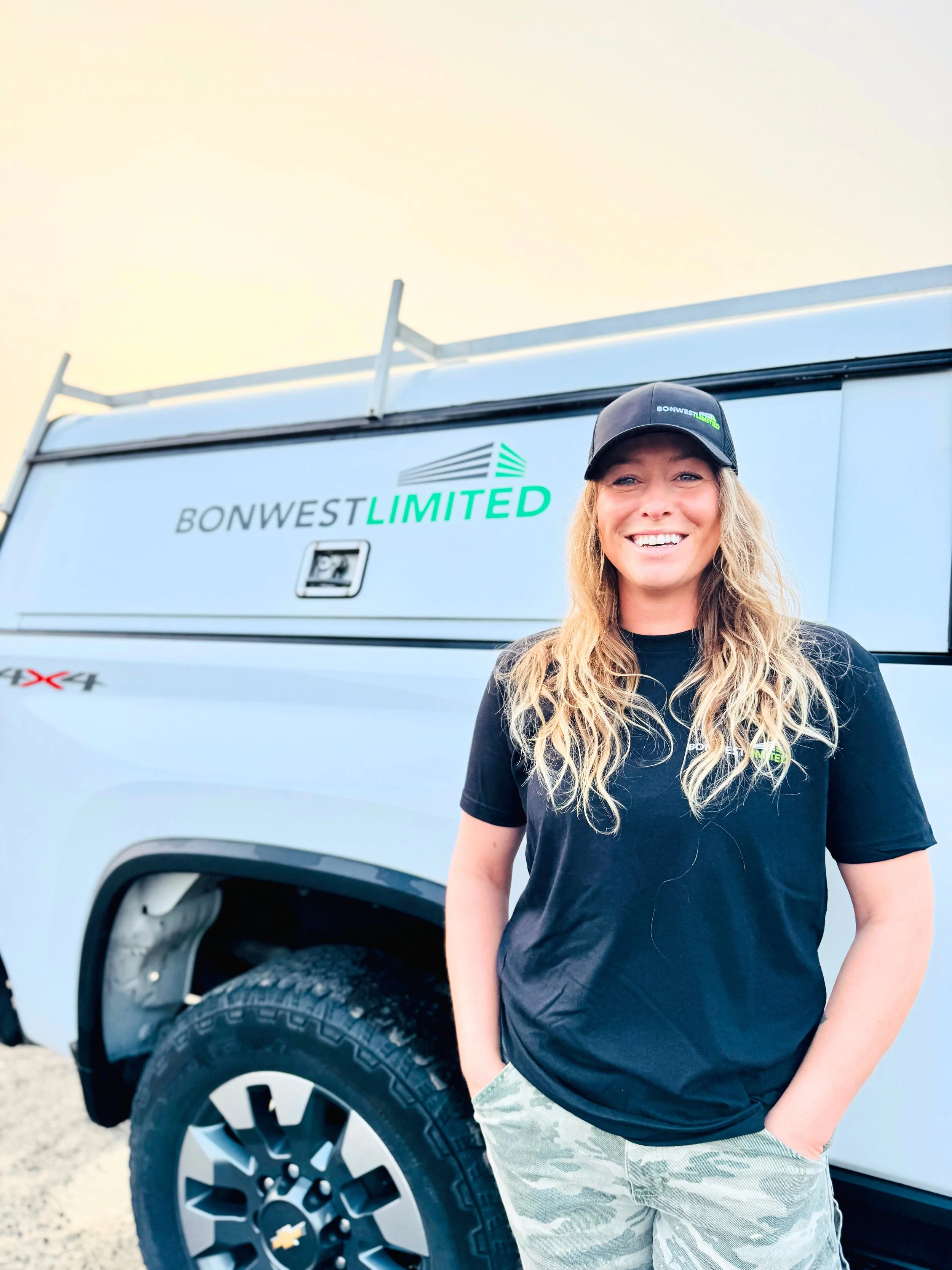 A woman with long blonde hair wearing a black cap, black t-shirt, and camouflage pants standing in front of a white BonwWst Limited company vehicle.