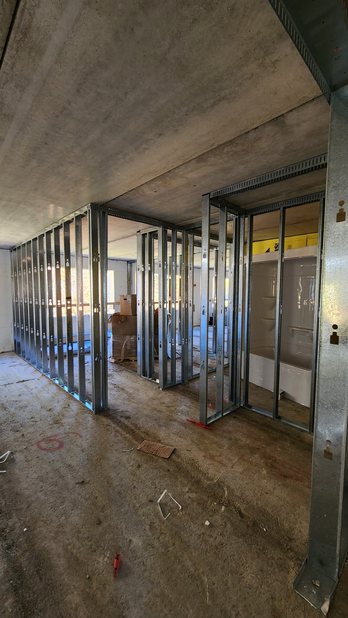 Interior of a building under construction with metal framing for walls and a bathtub installed, construction debris on the floor.