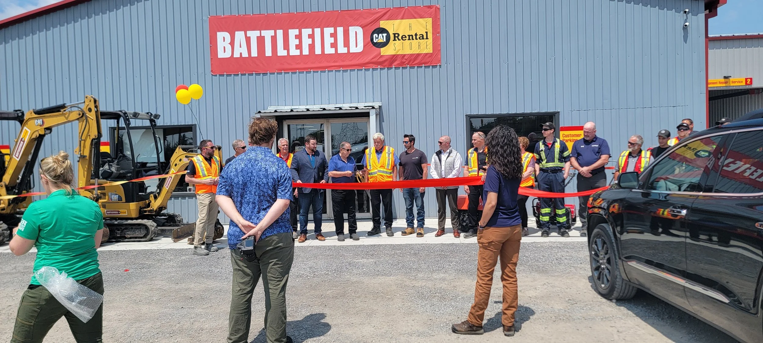 Ribbon-cutting ceremony outside a new construction equipment store with a large red "BATTLEFIELD" sign, around a group of people, some wearing safety vests, with construction machinery and cars nearby.
