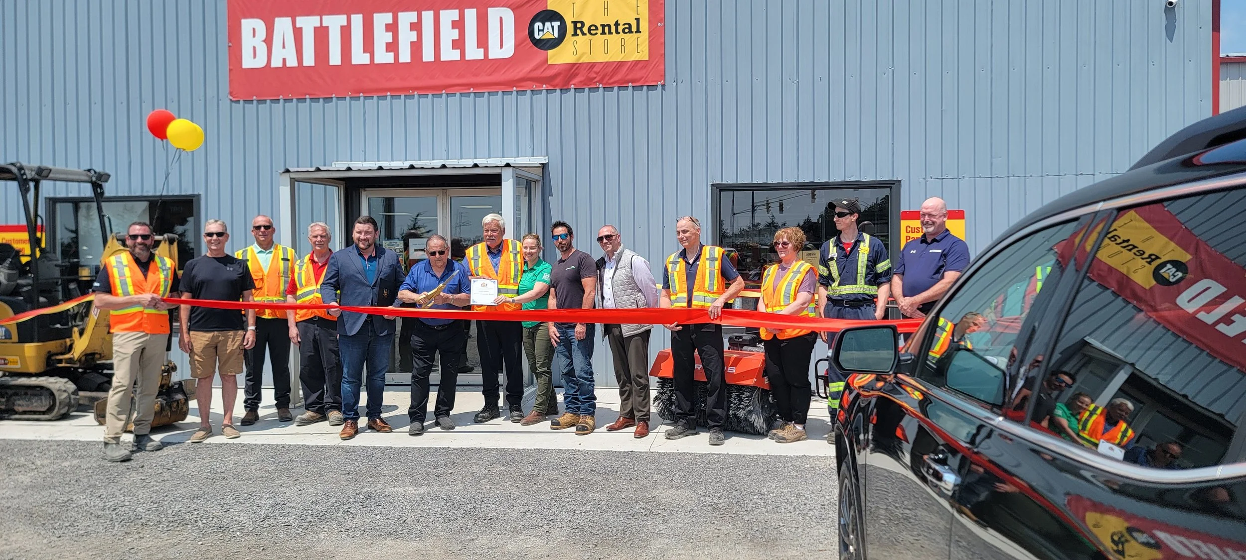 Group of people at a ribbon-cutting ceremony outside a building with a sign that reads "Battlefield" and "CAT Rental Store," with construction equipment and a black vehicle nearby.