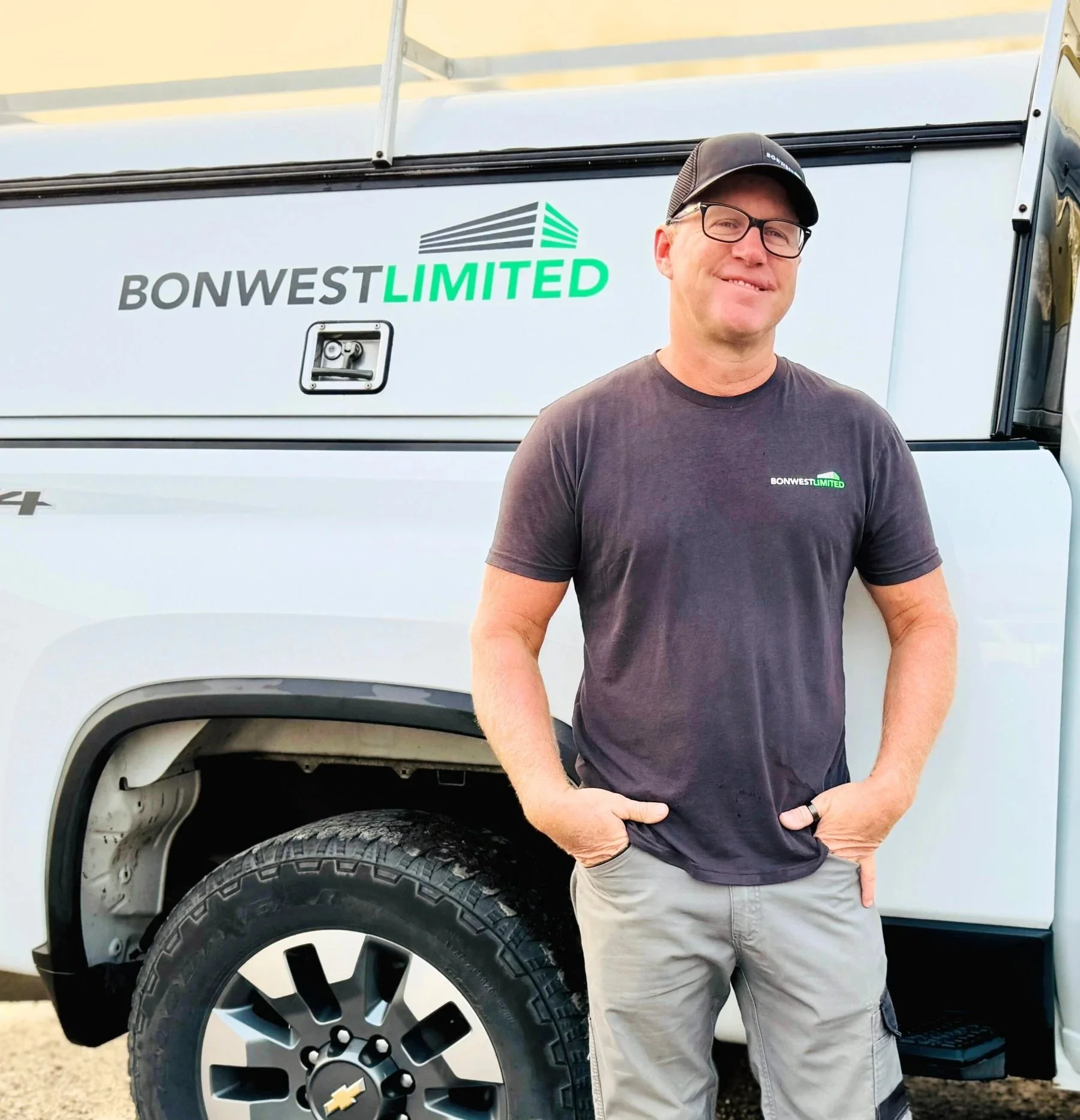 A man standing beside a white work truck with 'BONWEST LIMITED' logo, wearing a black cap, glasses, black T-shirt, and gray cargo pants, with hands in pockets.