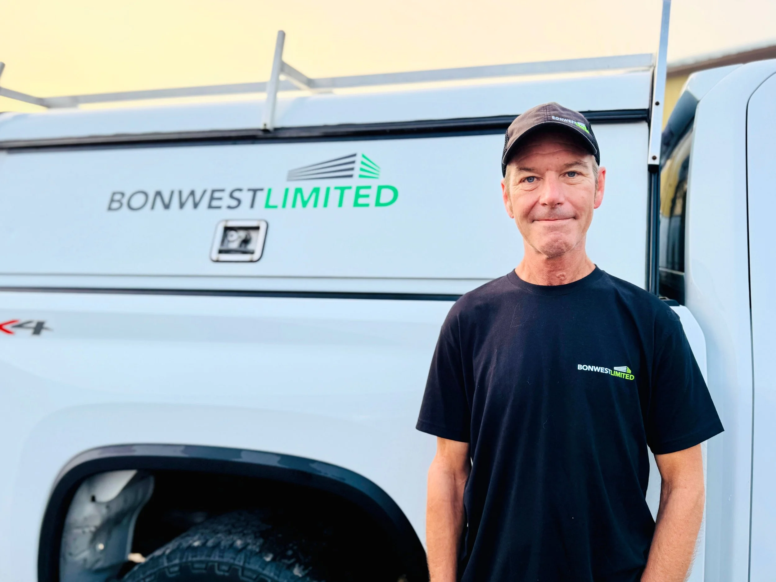 A man standing next to a white utility truck with the logo 'BONWEST LIMITED' on the side, wearing a black t-shirt and a black cap, smiling at the camera.