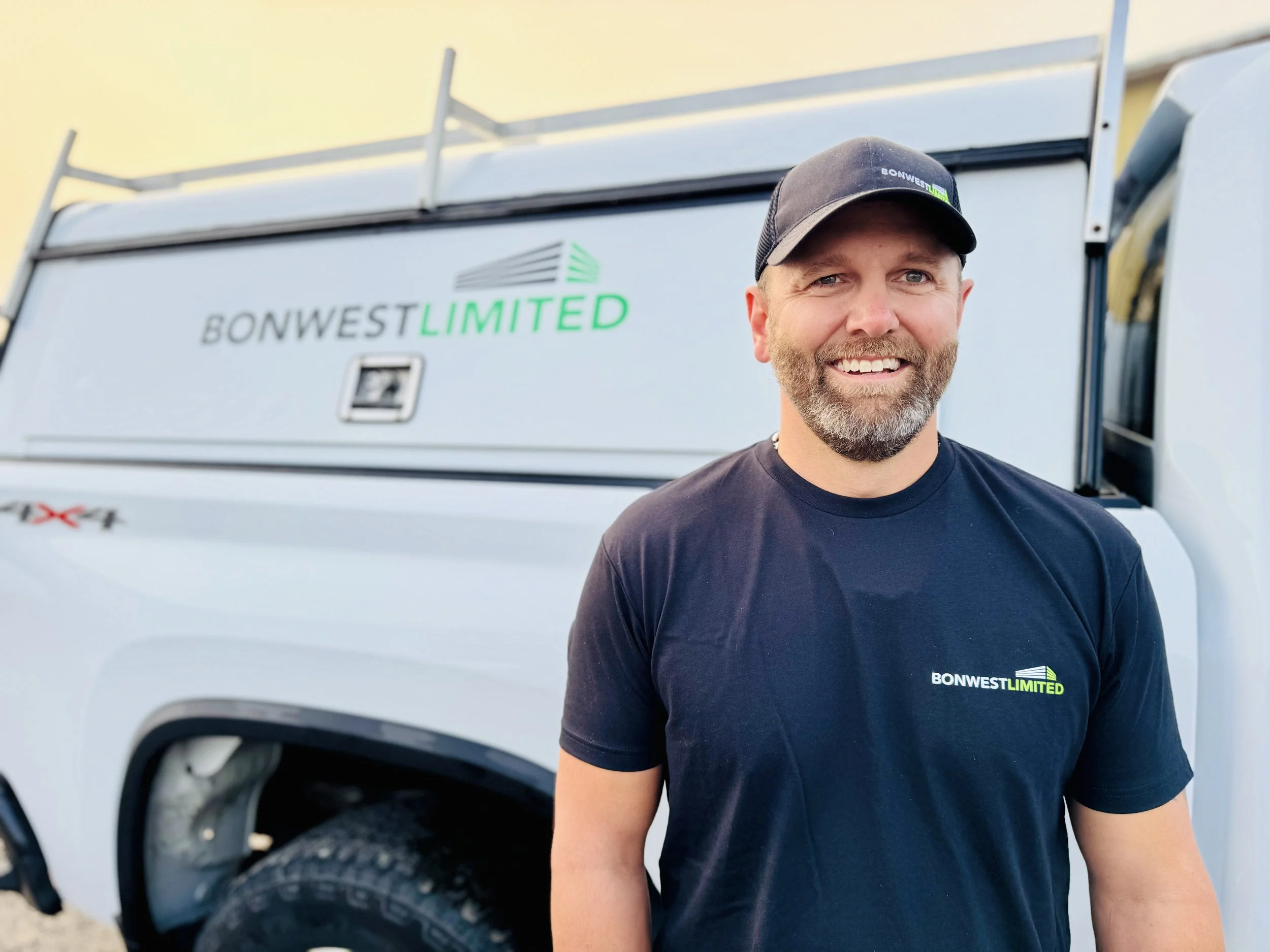 A man with a beard wearing a black cap and black t-shirt standing in front of a white company truck with "BONWEST LIMITED" logo.